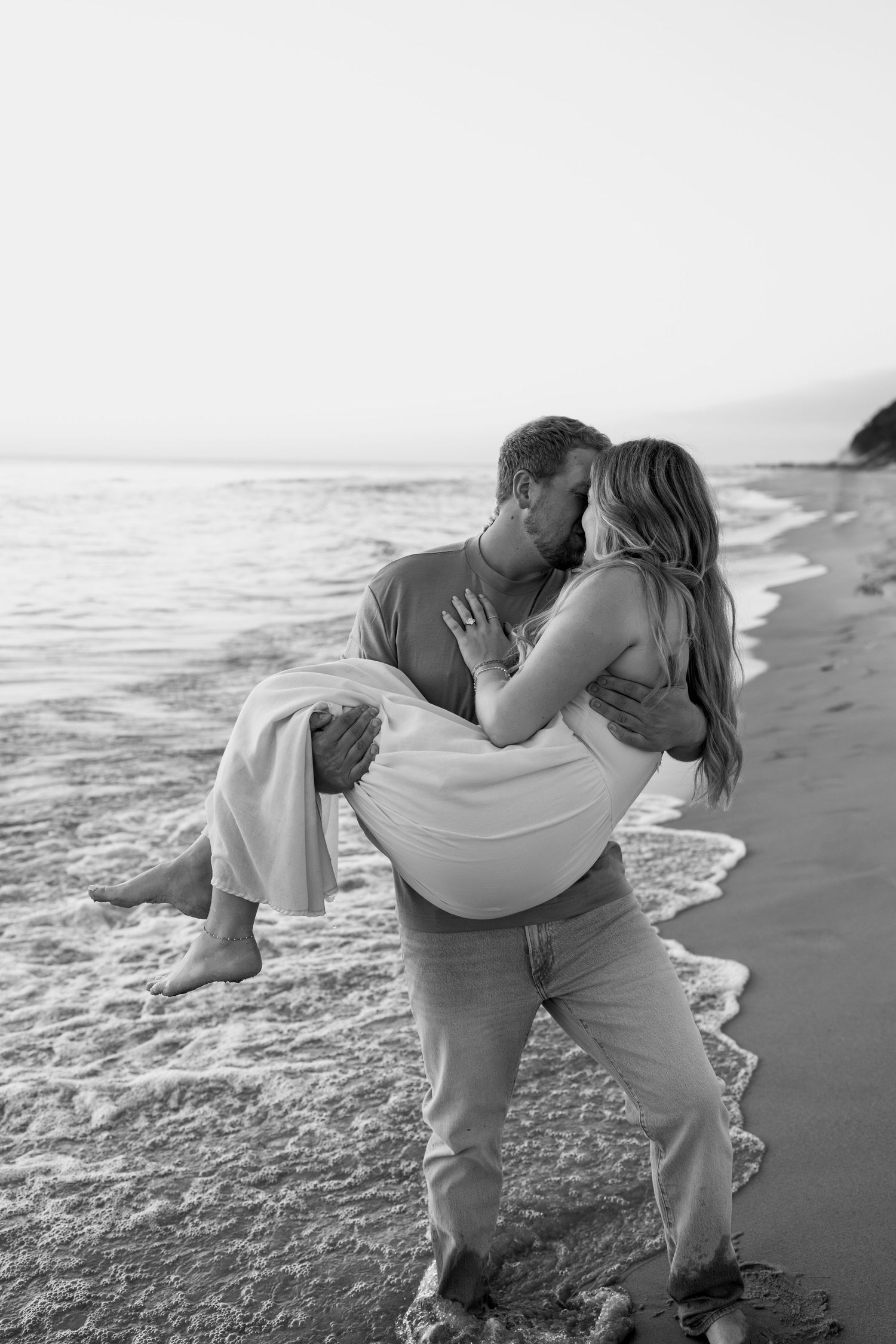 Black and white photo of a couple embracing on a sandy beach with ocean waves in the background.