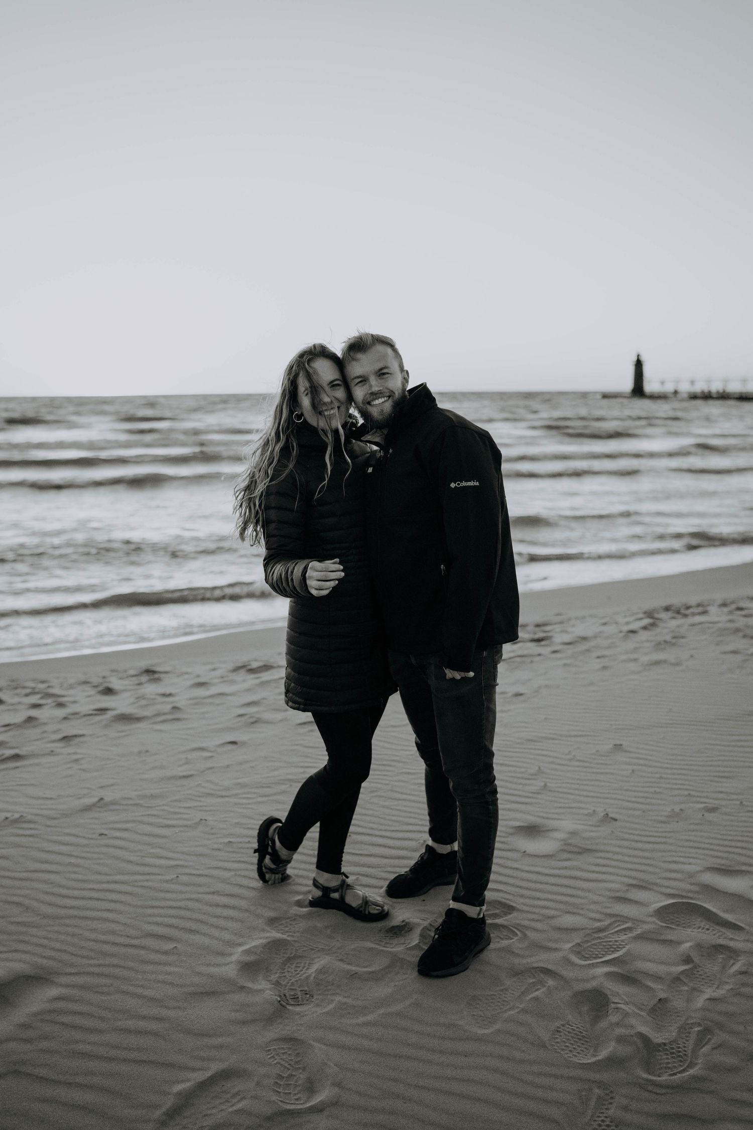 A couple embraces while walking along a moody black and white beach shoreline.