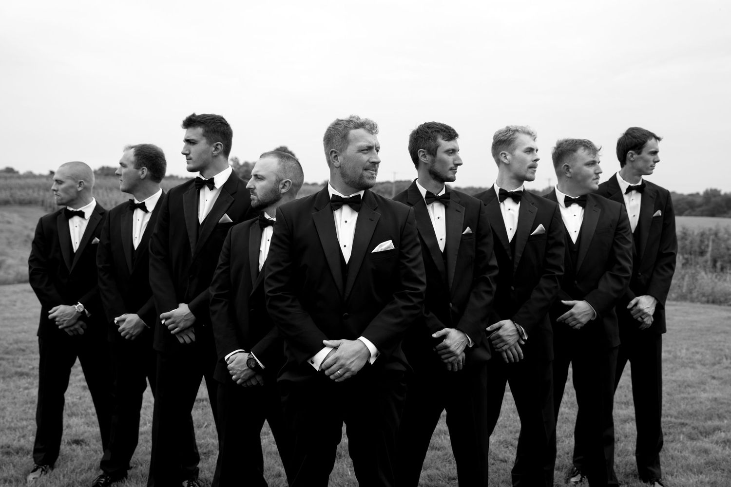 Black and white photo of a groom and groomsmen in tuxedos posing outdoors in a field.