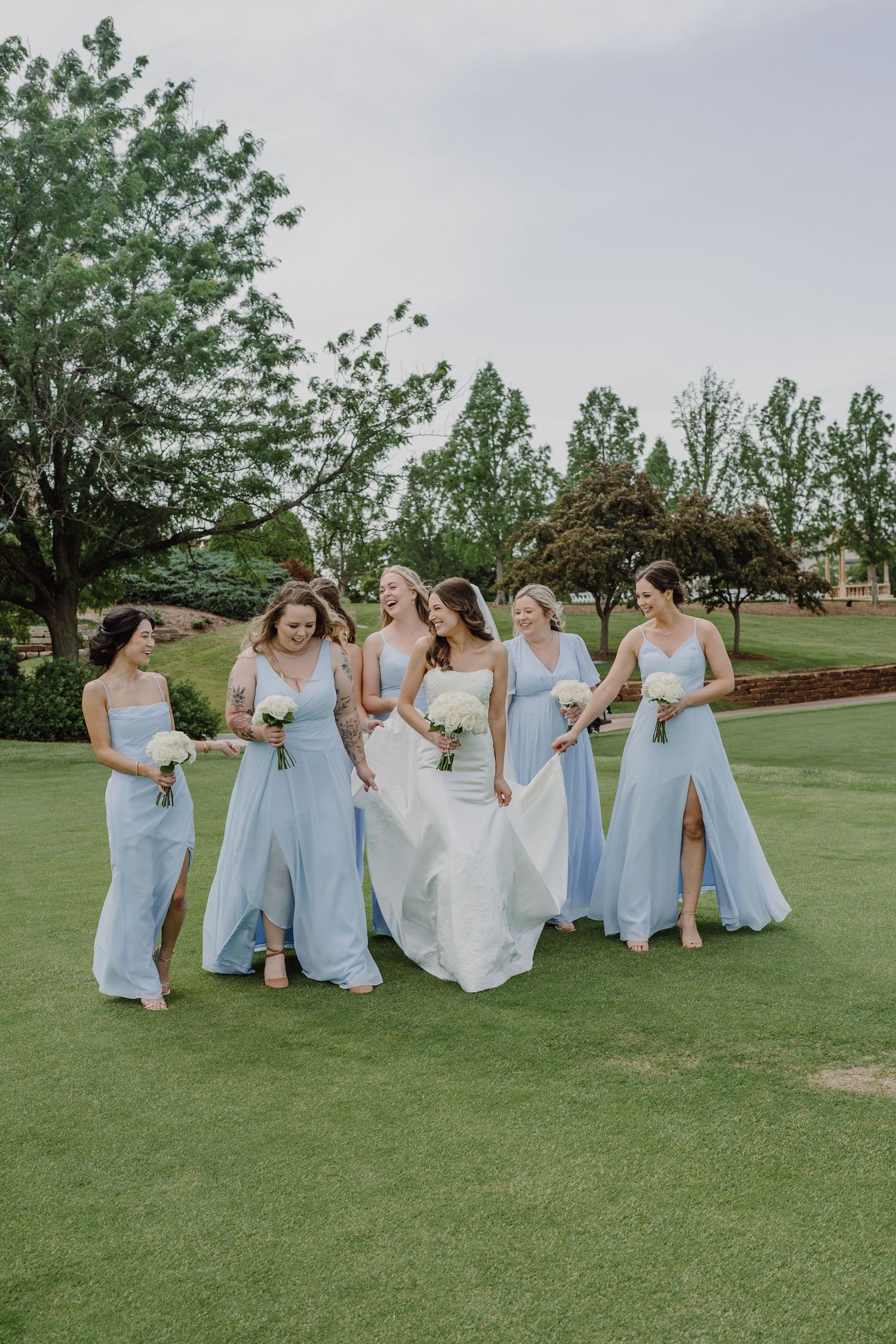 A bridal party in light blue bridesmaid dresses and white wedding gown pose playfully on a green lawn.
