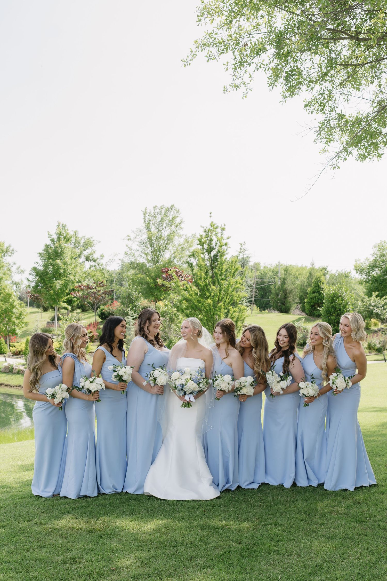 Bride in white gown with bridesmaids in light blue dresses holding white bouquets outdoors on green lawn.