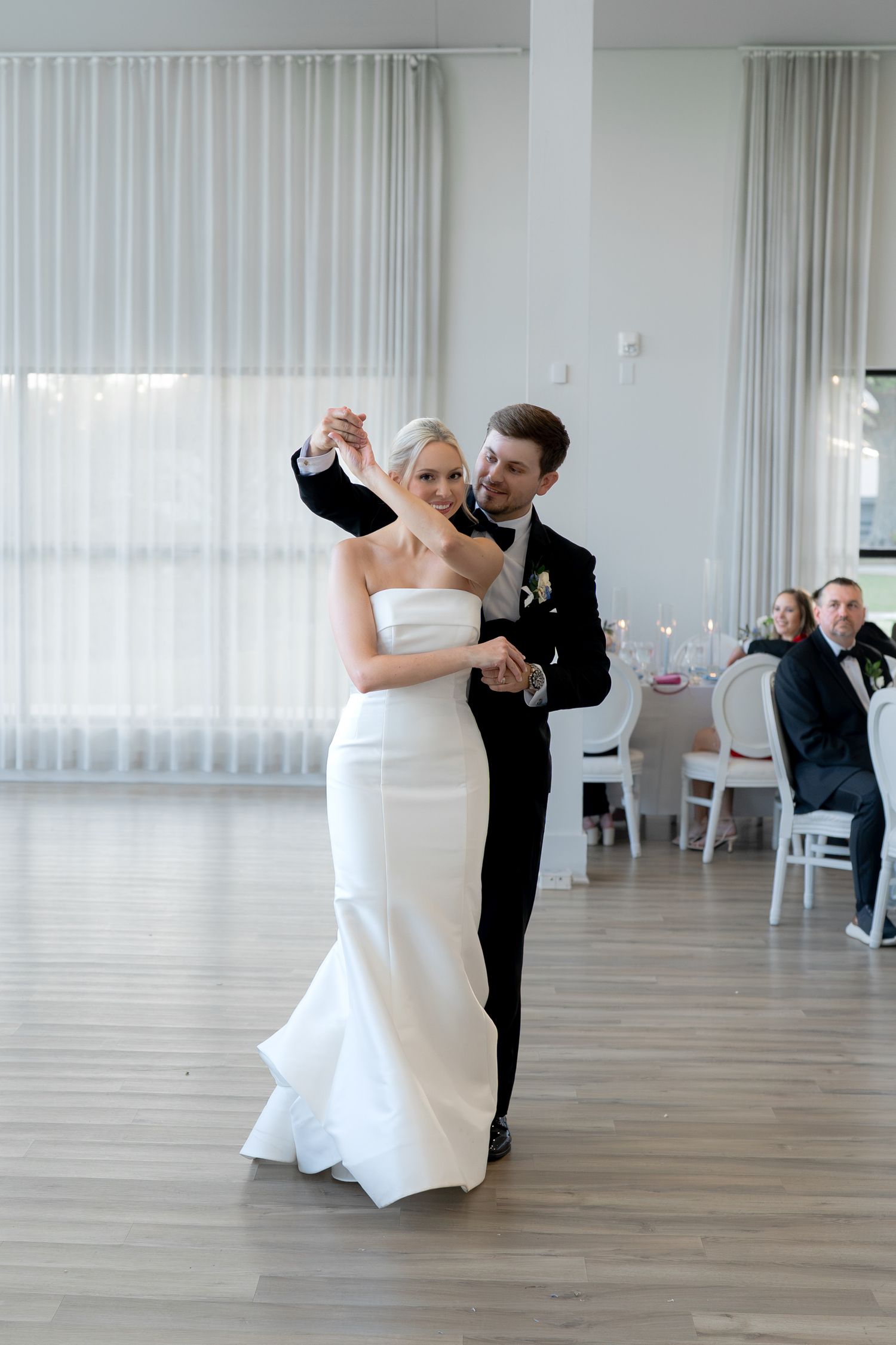 Bride and groom share their first dance in elegant white ballroom, groom spinning bride in white mermaid gown.