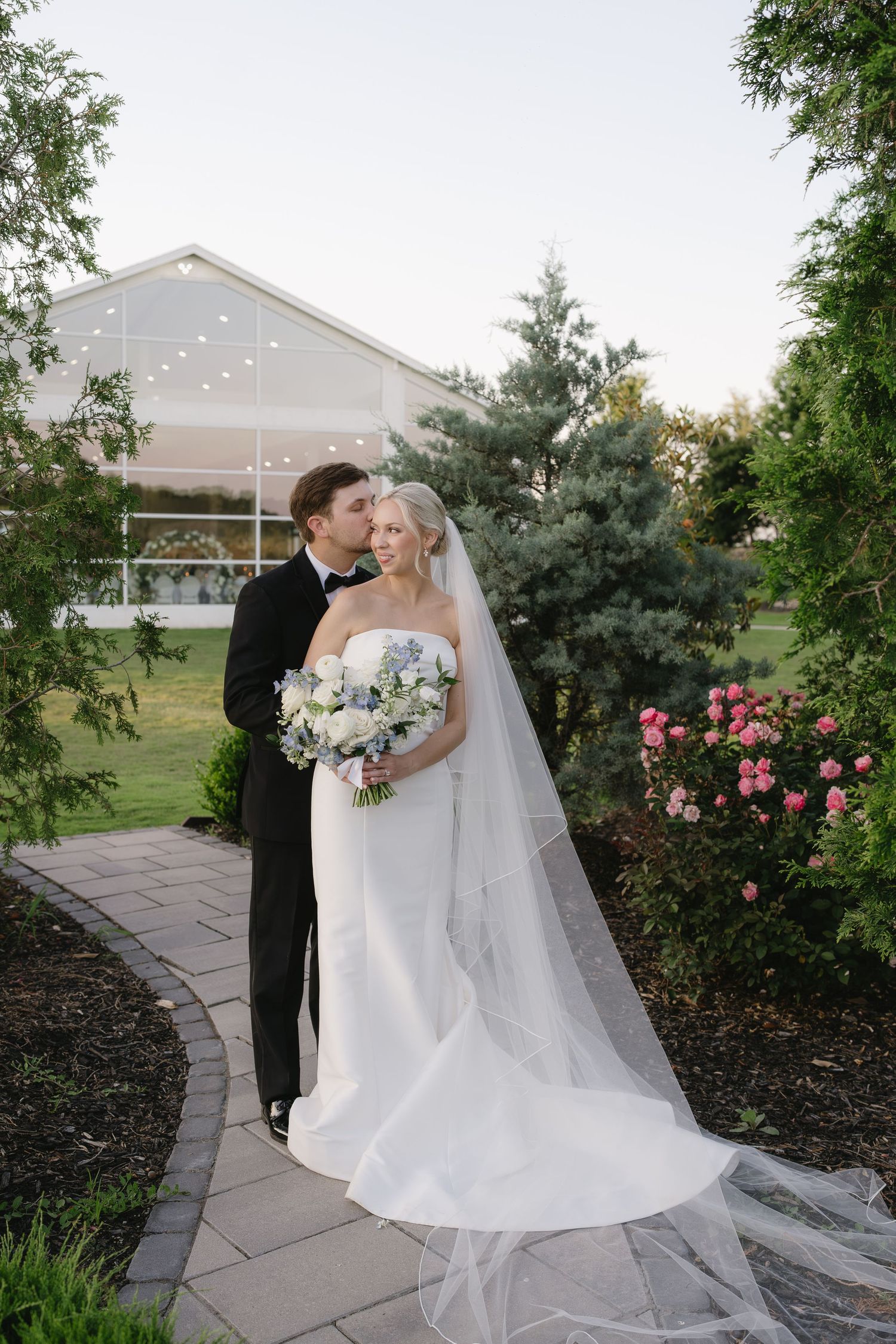 Bride and groom share a romantic kiss on a garden path, with a white wedding venue and blooming flowers in the background.