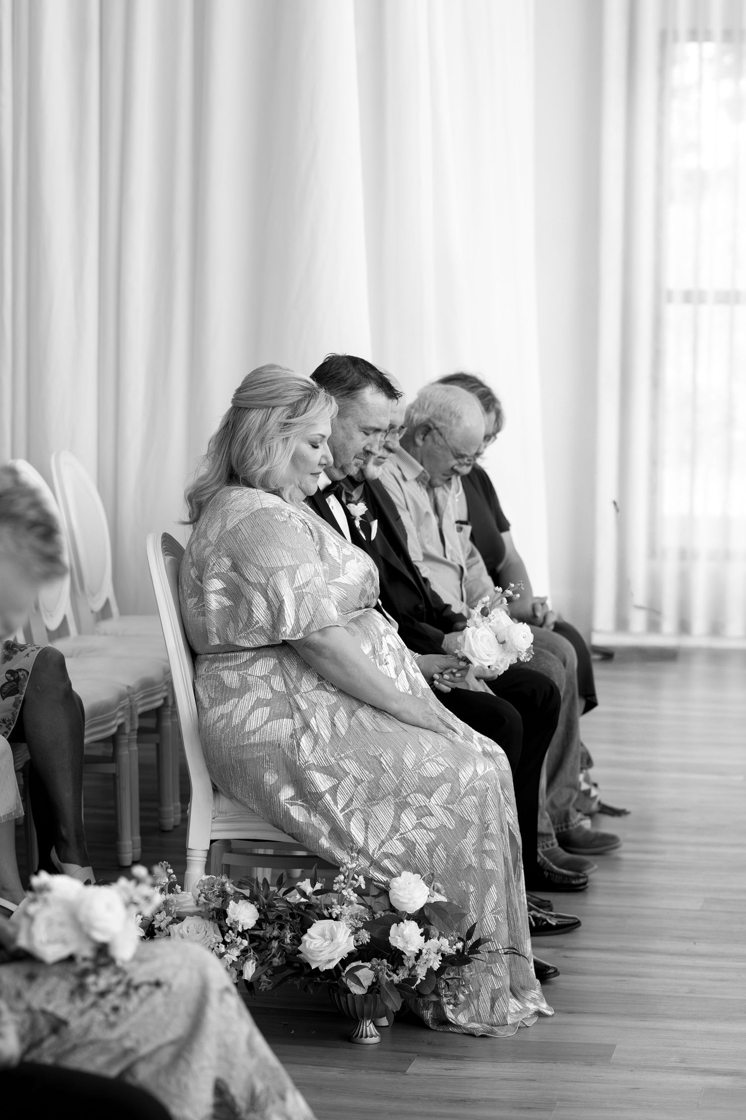 Black and white photo of wedding guests seated in chairs during a ceremony, with floral arrangements on the floor.