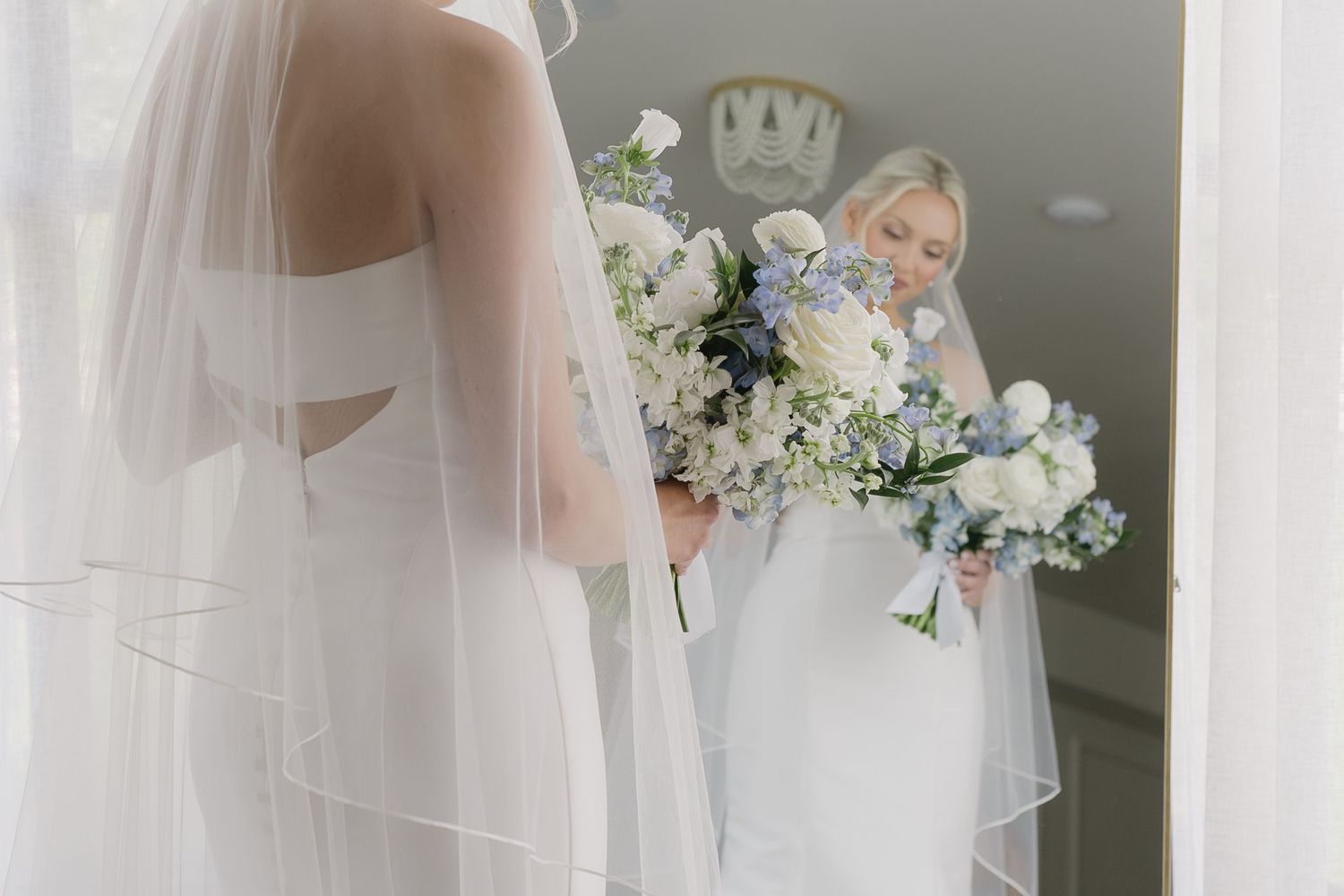 Bride in white gown and veil holds blue and white bouquet, reflected in mirror during wedding preparations.