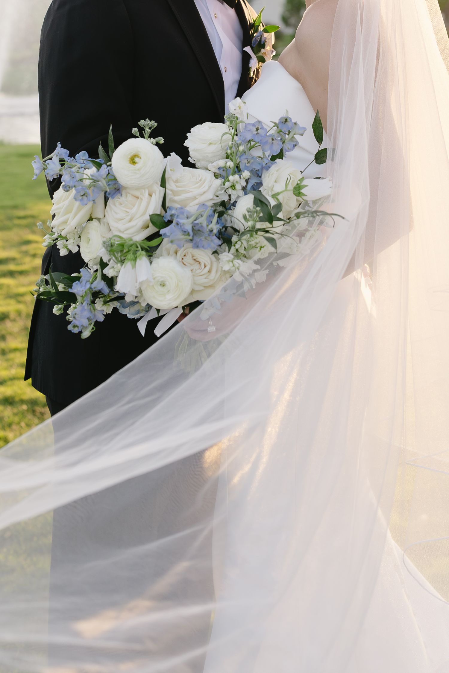 Bride and groom holding a white and blue floral wedding bouquet with roses, with veil flowing in the breeze.