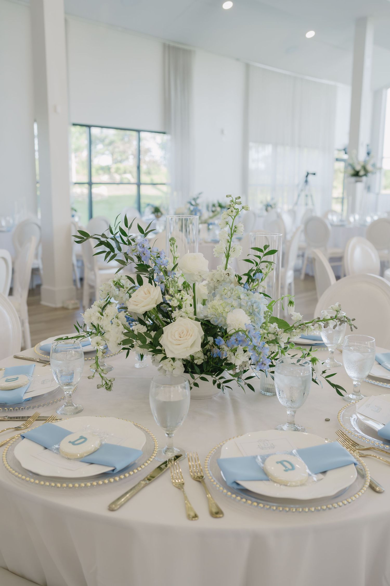 Elegant wedding reception table with blue and white floral centerpiece, gold cutlery, pearl chargers, and light blue napkins.