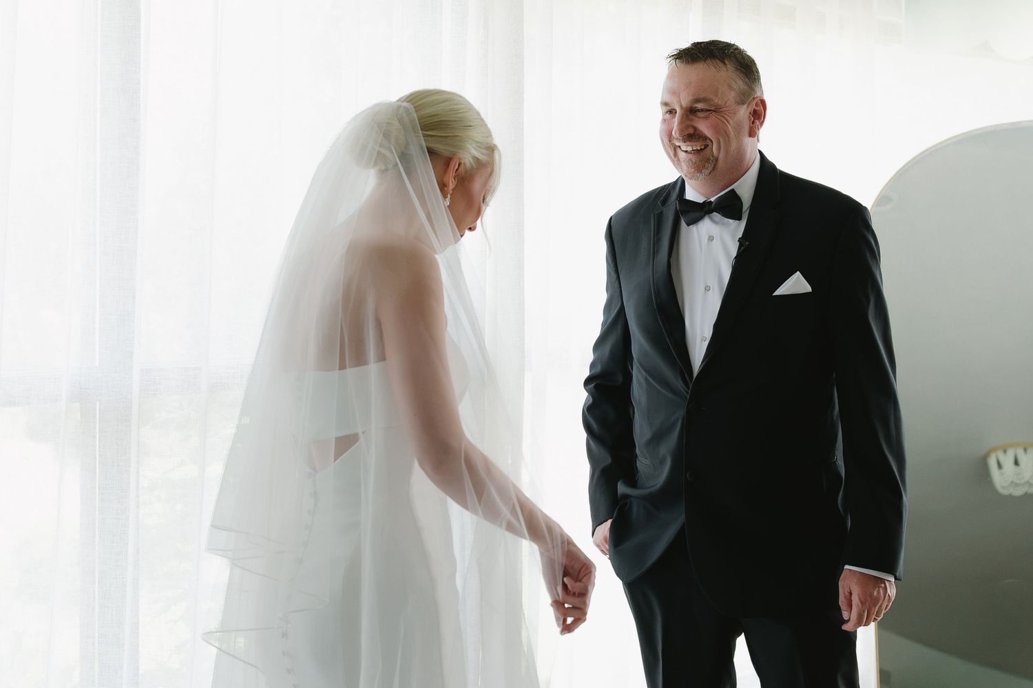 Bride in white gown and veil shares a joyful first look moment with groom in black tuxedo.