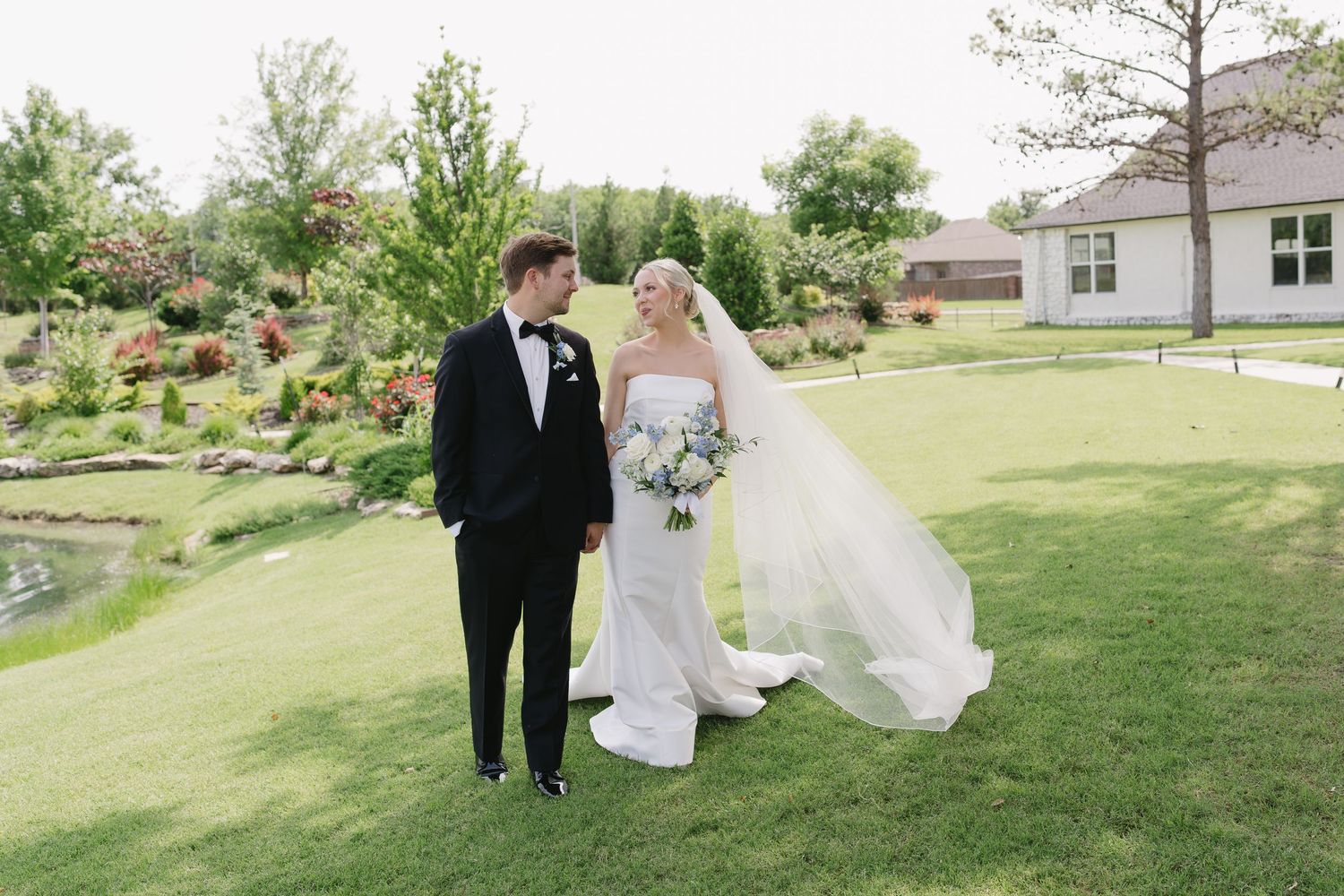 Bride and groom walking on lush green lawn at outdoor wedding venue with garden and pond backdrop.