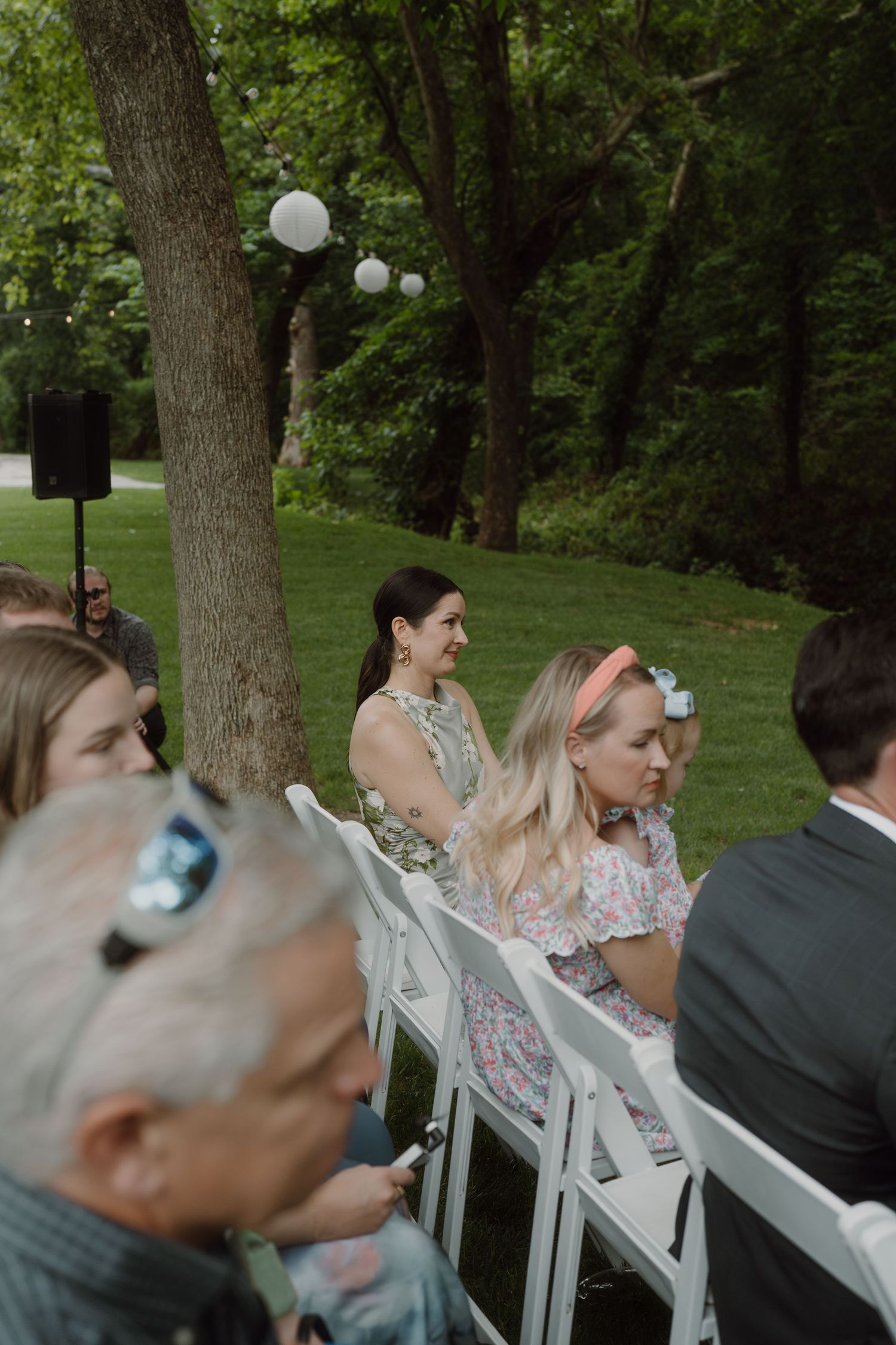 Wedding guests seated in white chairs beneath tall trees during an outdoor ceremony.