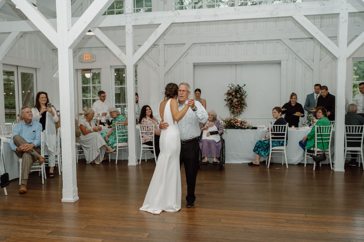 Couple sharing their first dance in a rustic white barn reception venue with guests seated around them.