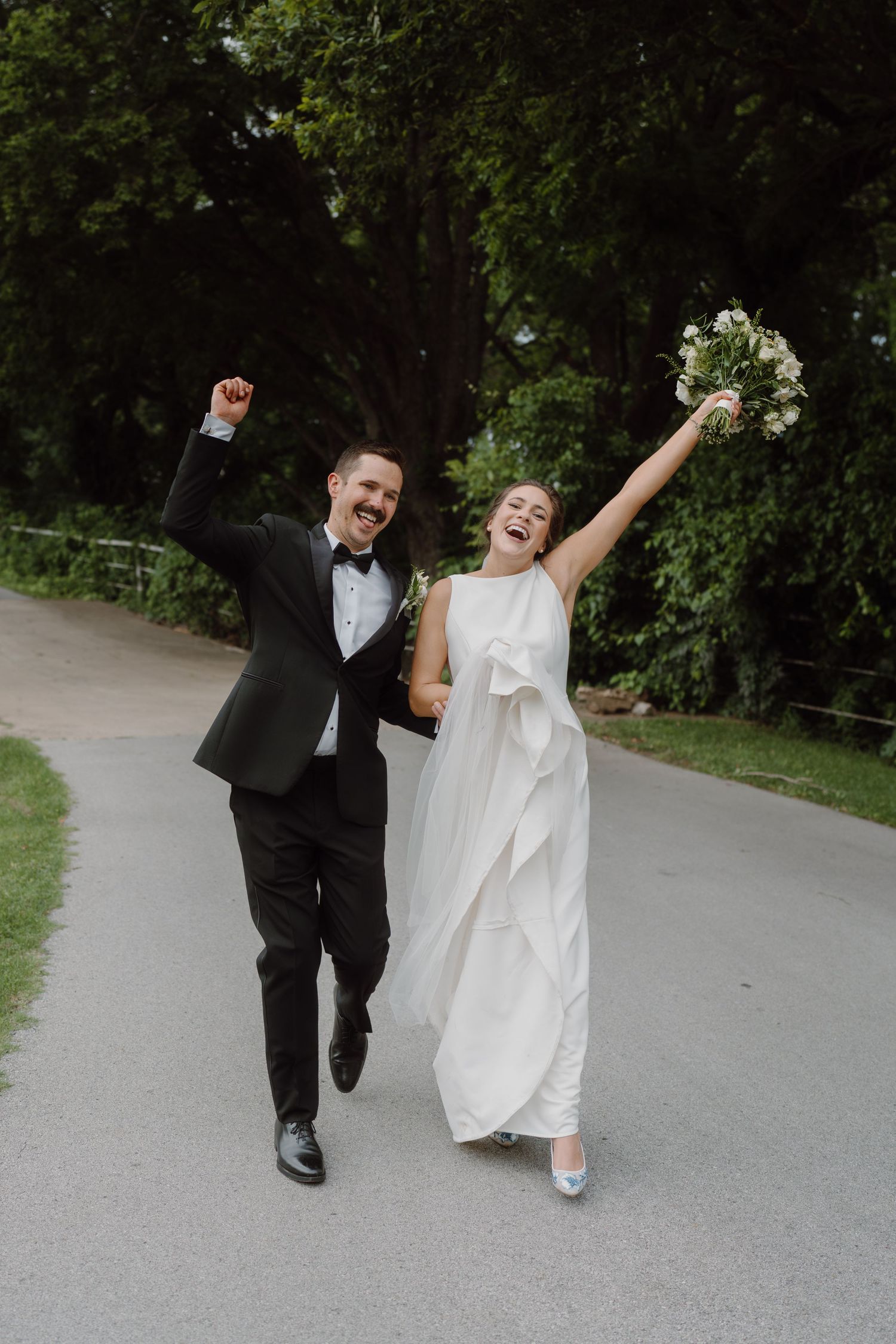 Joyful wedding couple celebrates while walking down a paved path surrounded by trees.