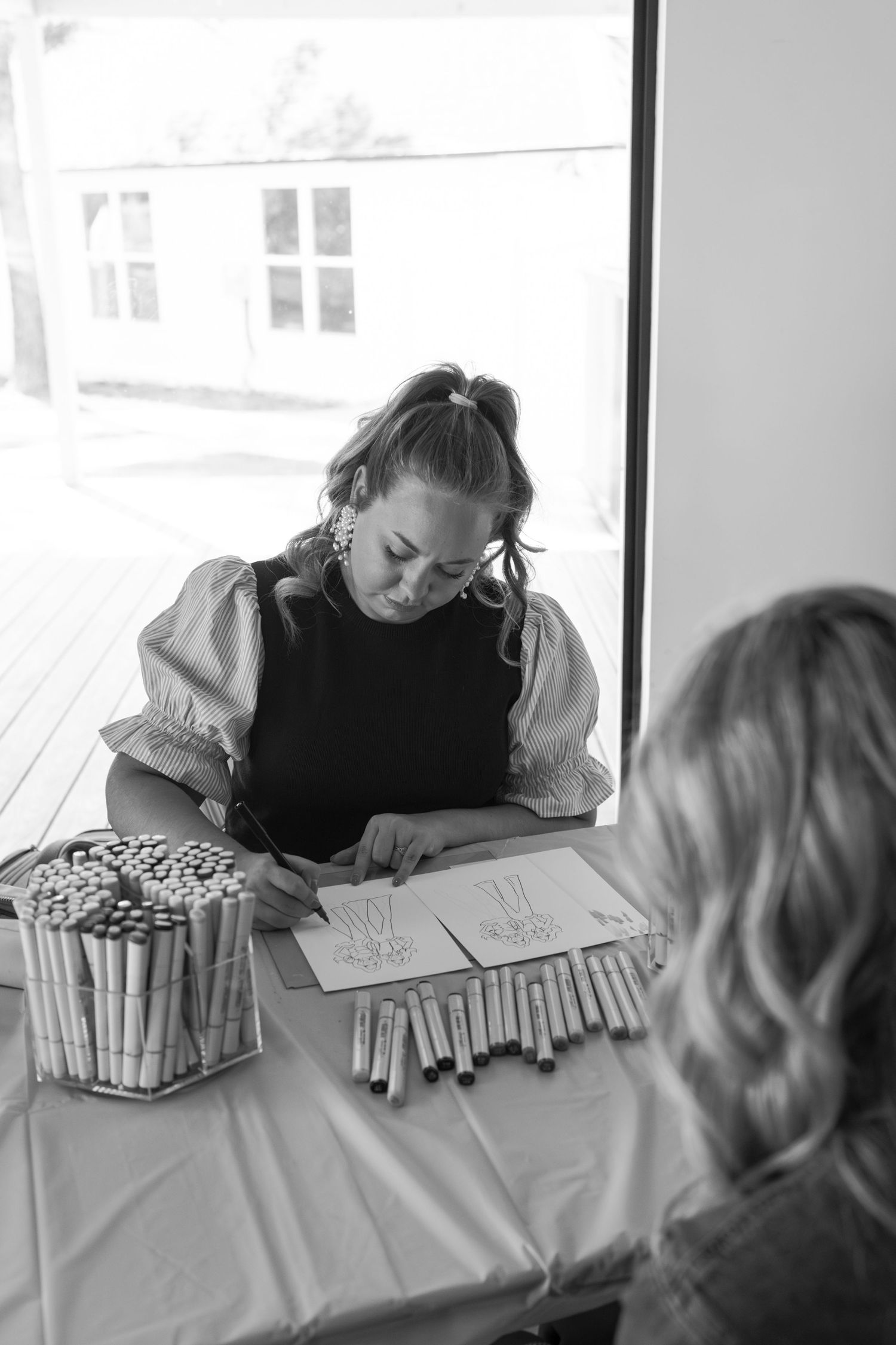 A person writing or drawing on paper while sitting at a table in a bright room.