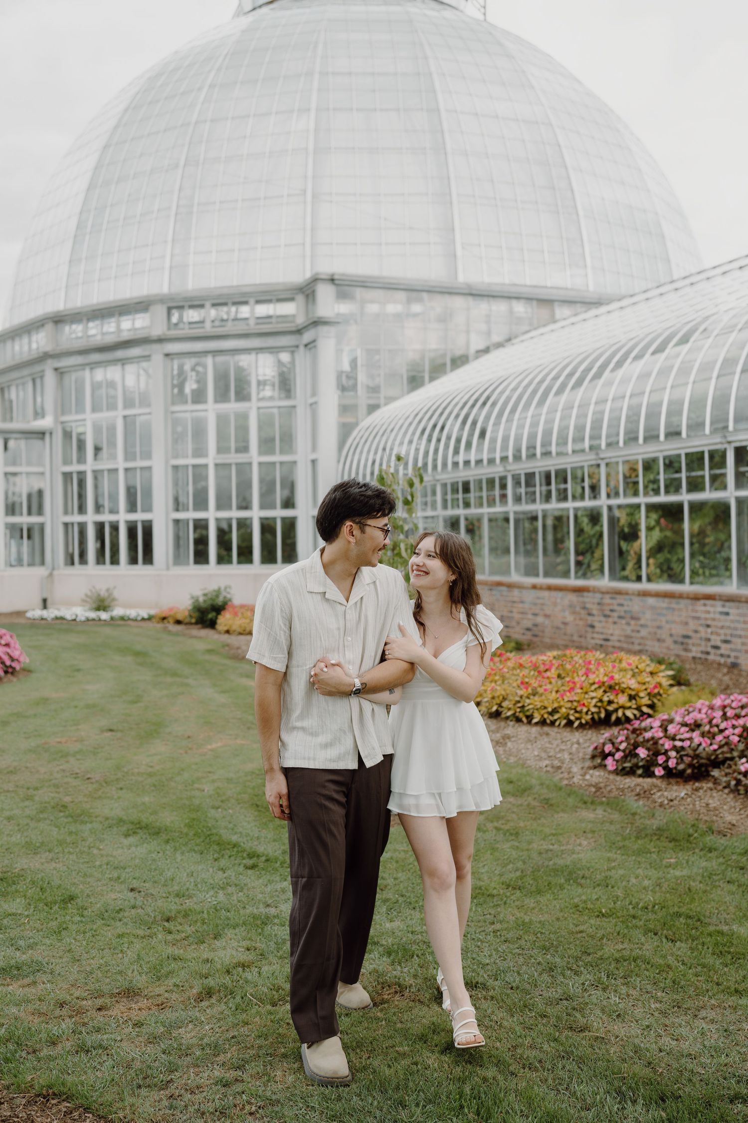 Two people in light summer clothing share a moment near a conservatory with colorful gardens.