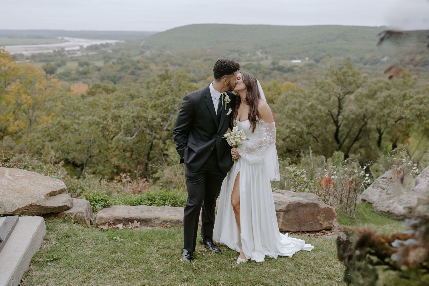 A scenic wedding photoshoot on a hilltop overlooking a vast natural landscape with rolling hills and greenery.