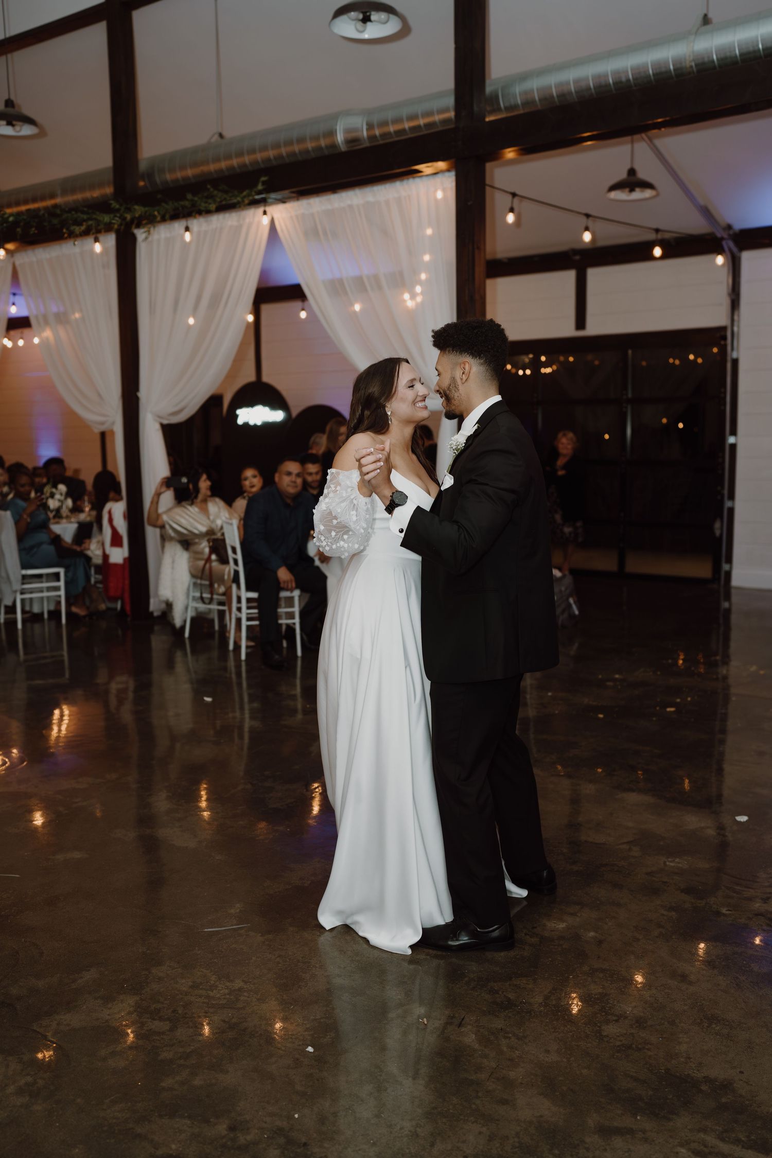 A romantic dance moment captured in a modern wedding venue with white drapes and candlelit floors.
