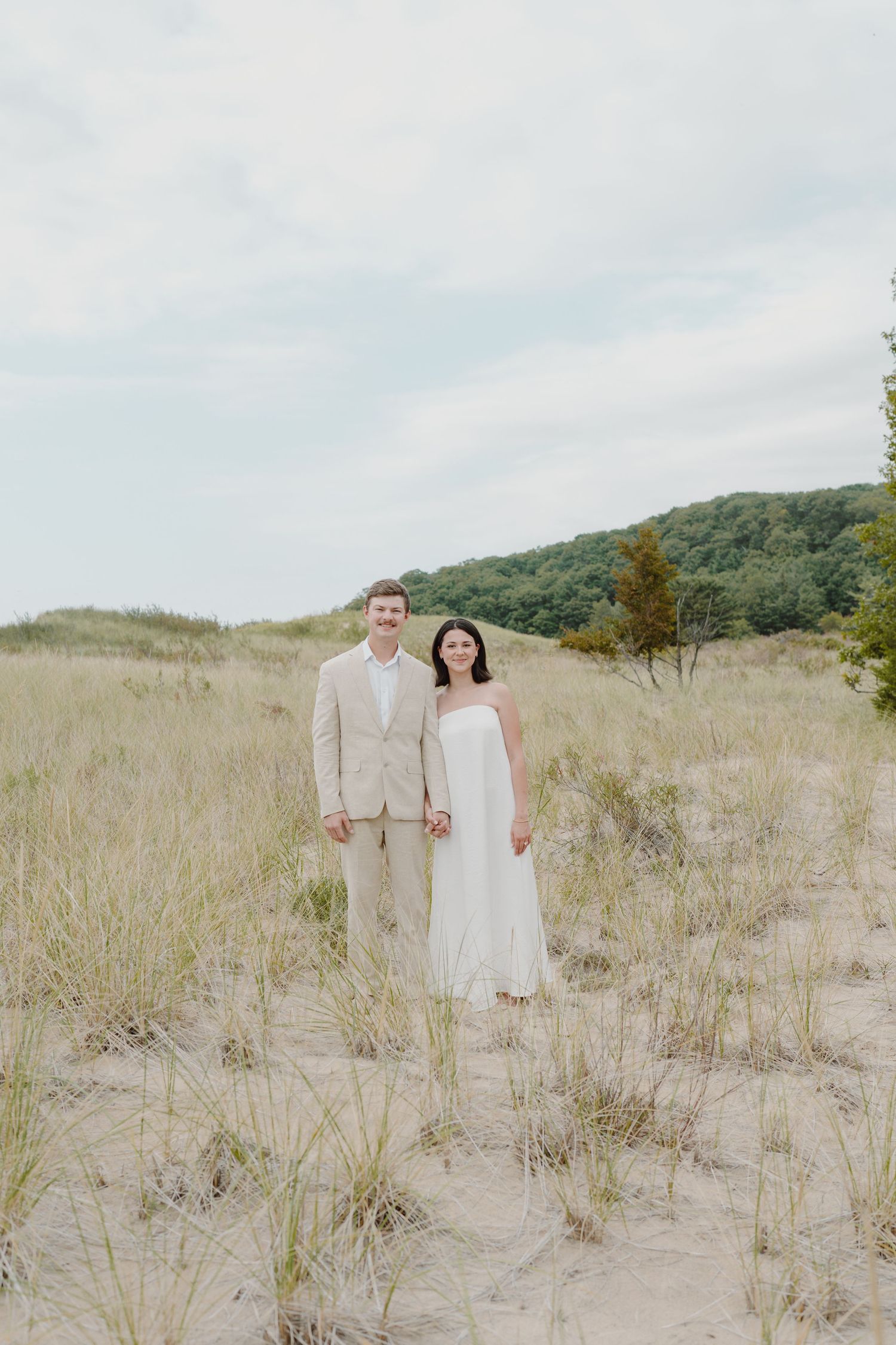 A couple stands together in a scenic dune landscape with trees and hills in the background.