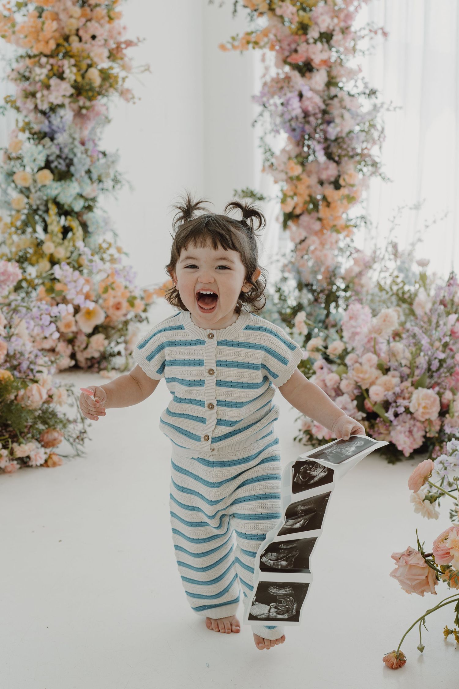 Child in striped outfit jumping joyfully in front of floral backdrop during photo session.