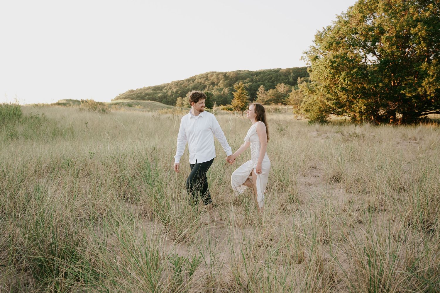 Two figures walk together through a serene meadow landscape with hills in the distance.