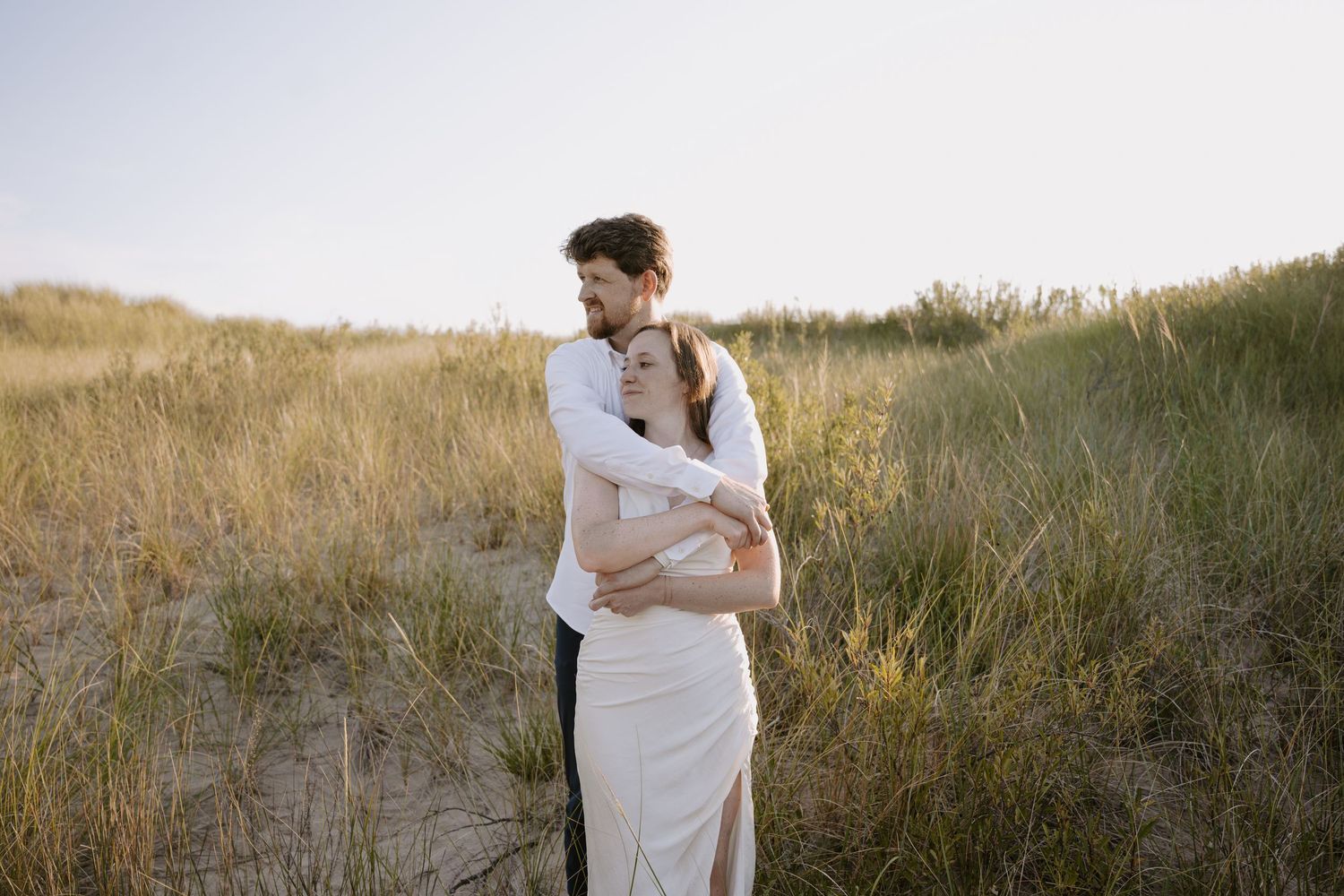 Two people share an intimate embrace on a coastal dune landscape.