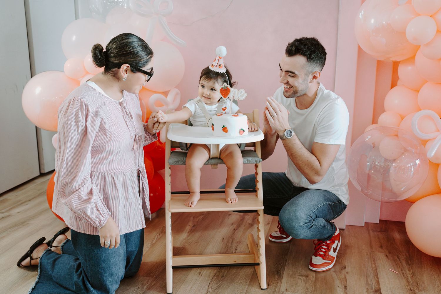 Birthday party setup featuring pink balloon arch and white wooden high chair.