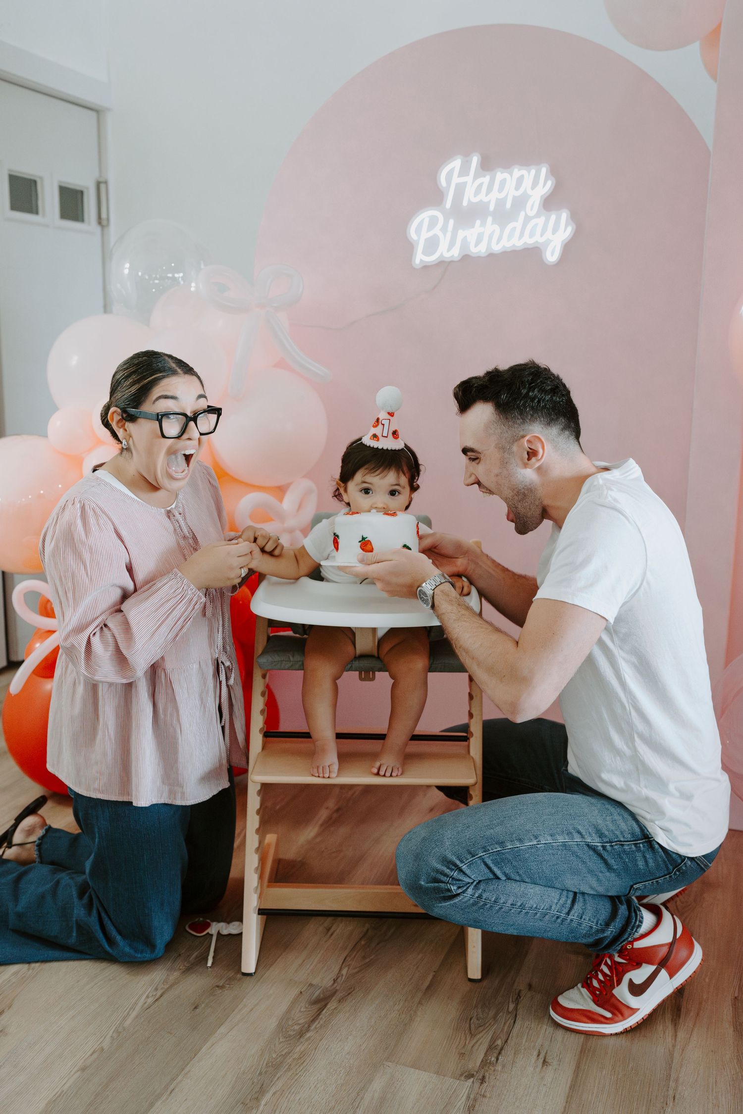 Parents sharing laughs with baby during birthday celebration with pink balloon backdrop.