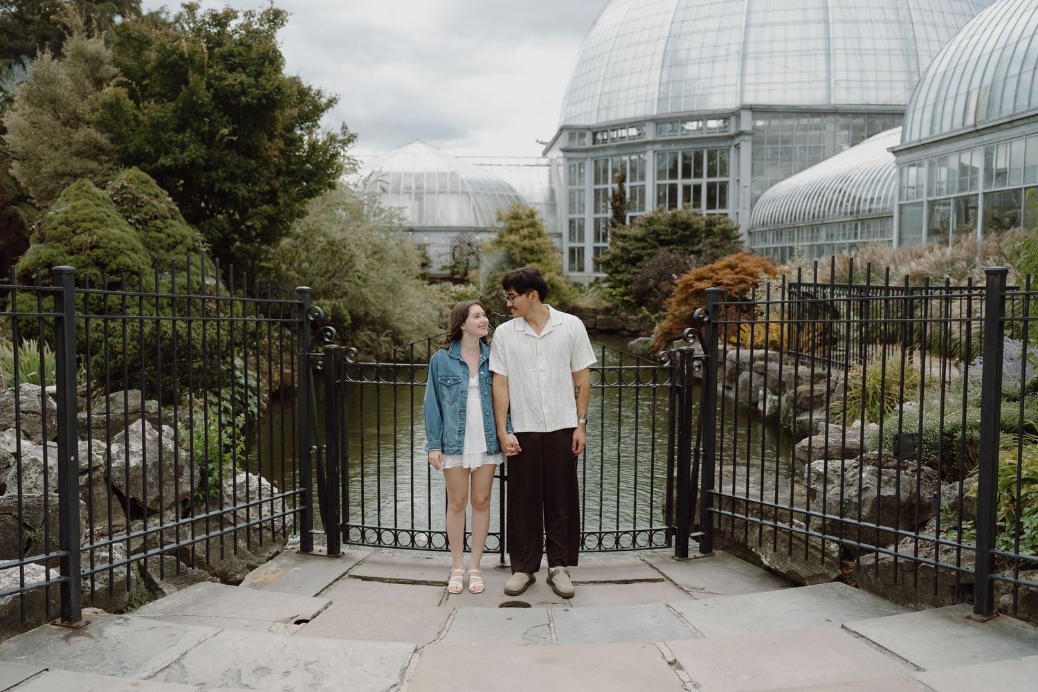Two people pose on pathway with ornate black metal railings and glass conservatory dome in background.