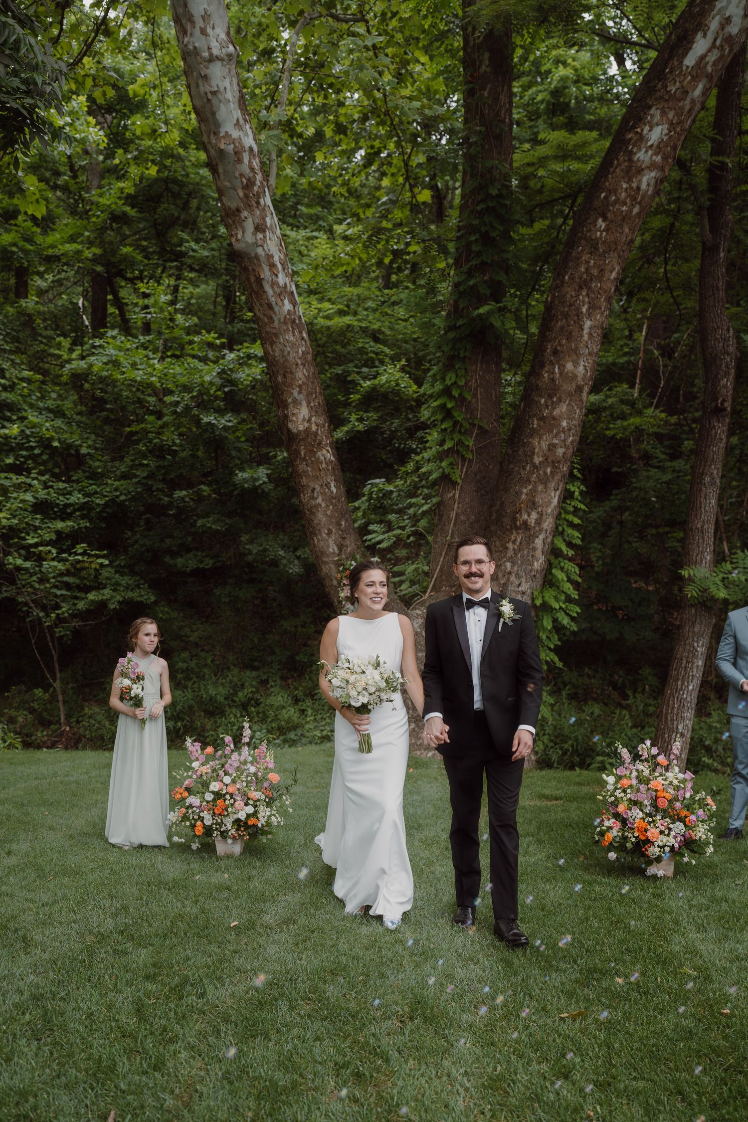 Wedding processional in a wooded area with bright floral arrangements and guests looking on.