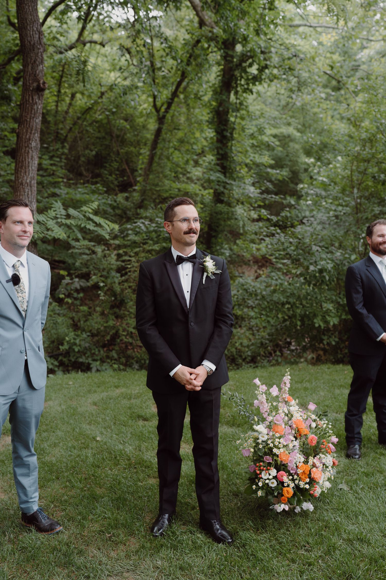 Groomsmen in formal attire stand near floral arrangements in a wooded garden setting.