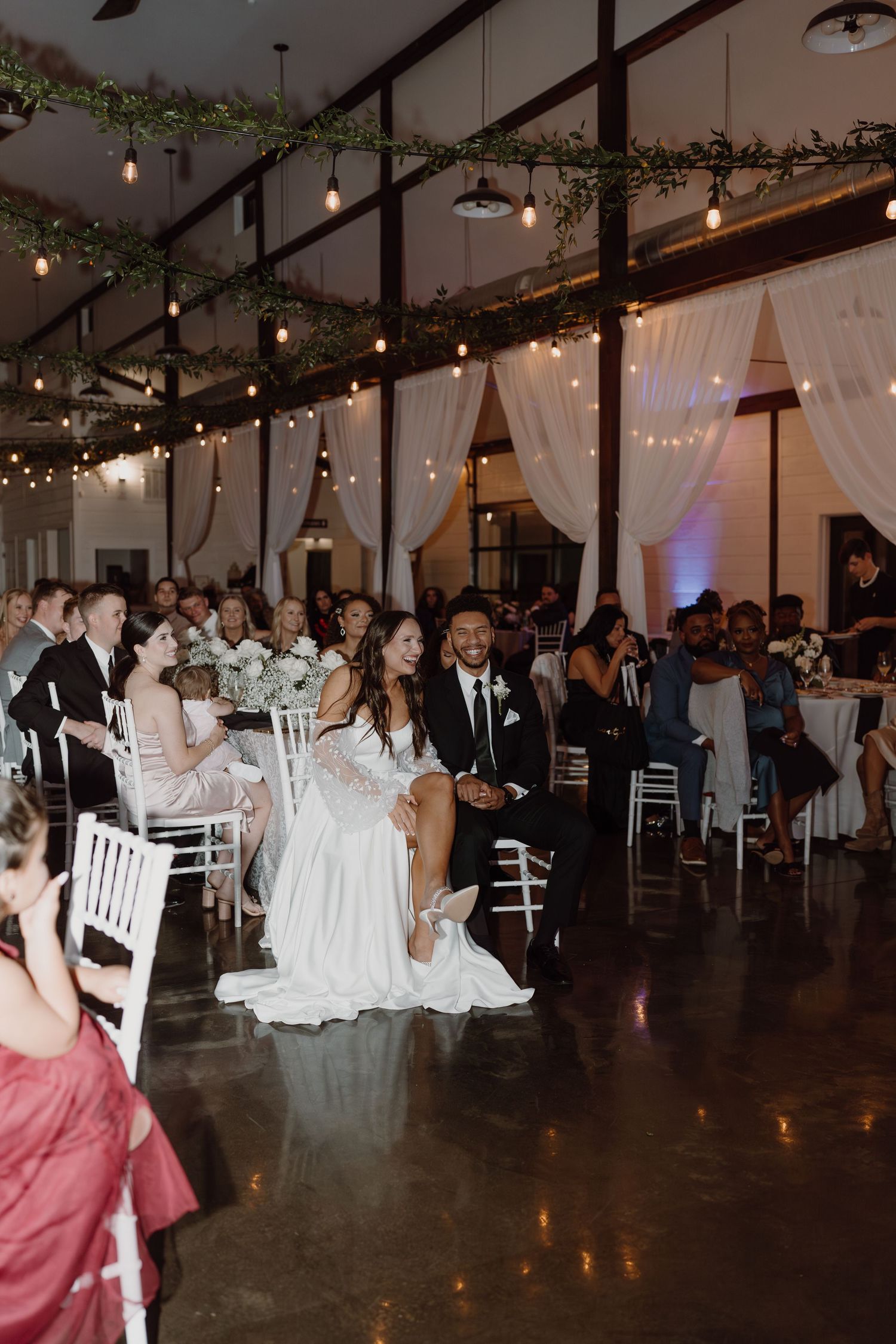 Wedding reception guests dancing and celebrating in an elegantly decorated venue with string lights and white drapes.