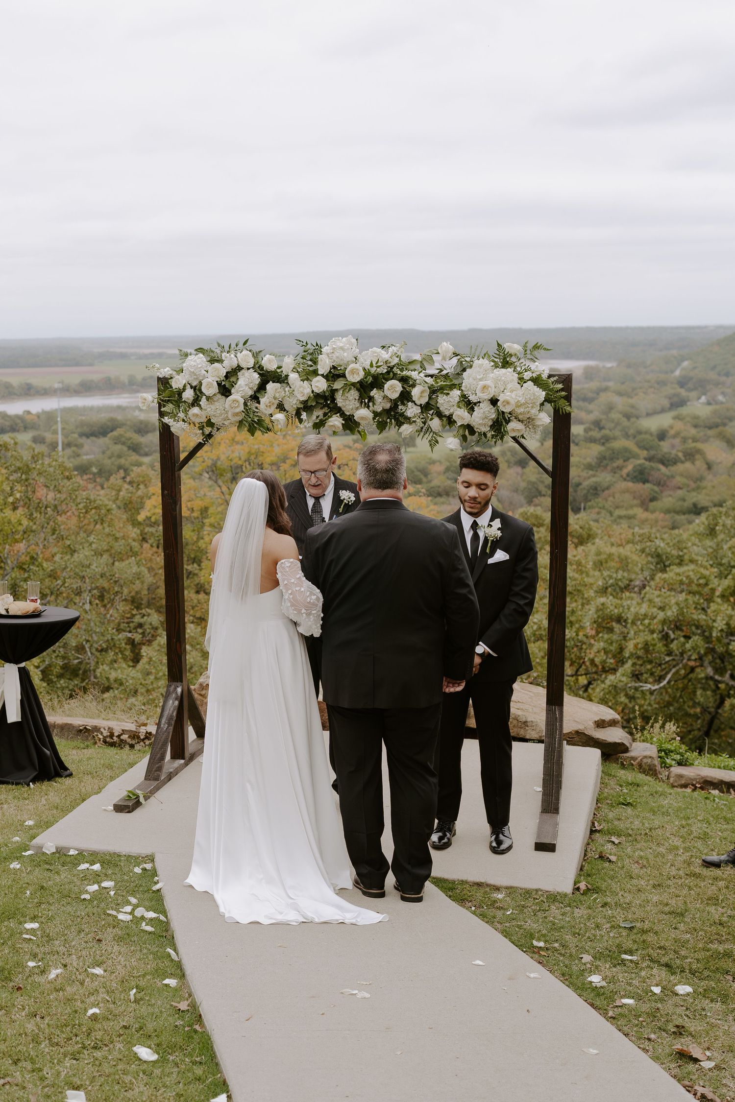 Wedding ceremony taking place under a wooden arch decorated with white flowers on a scenic overlook.