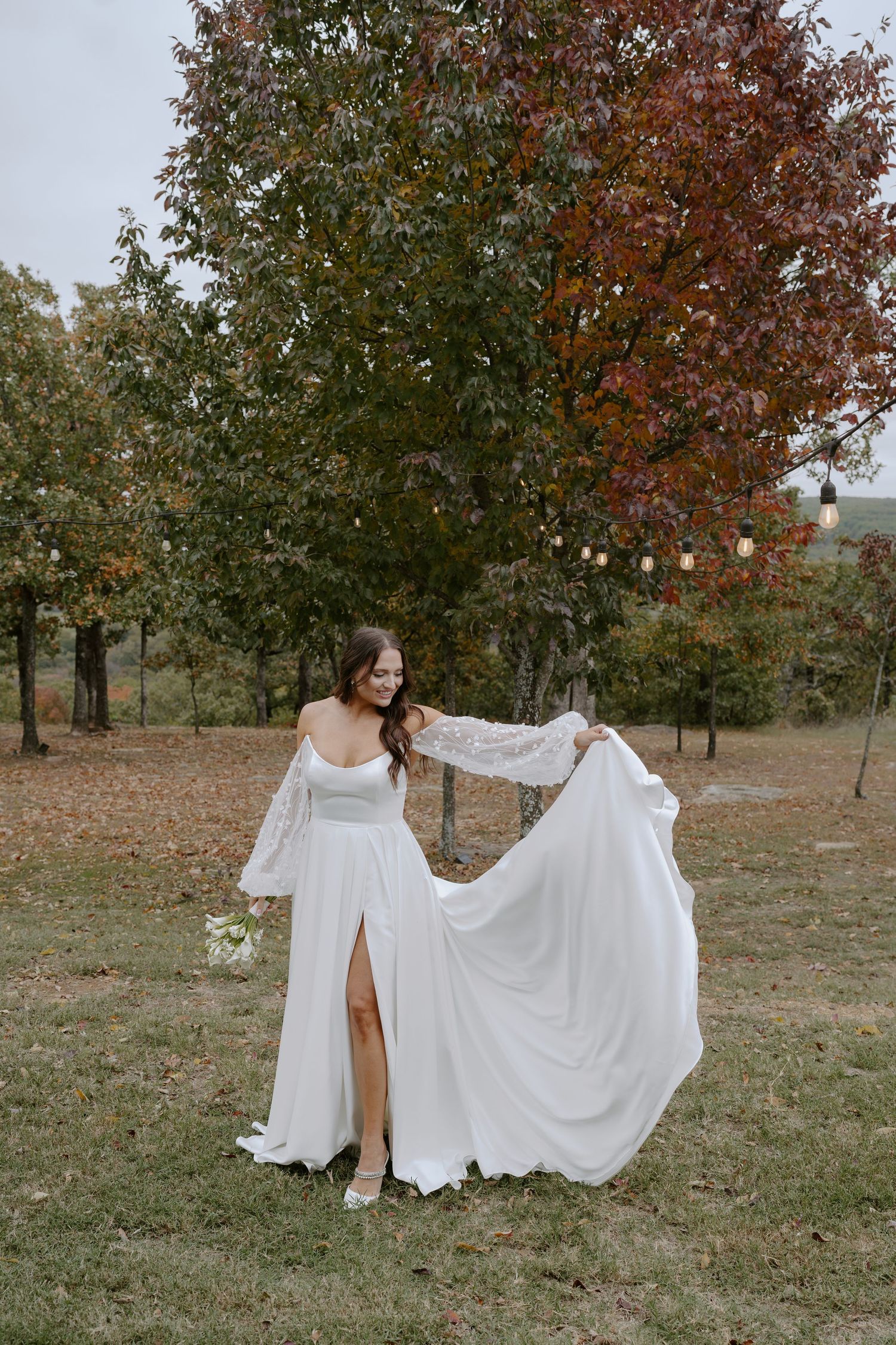 A wedding dress with long lace sleeves and a flowing skirt is displayed in an autumn outdoor setting with string lights.
