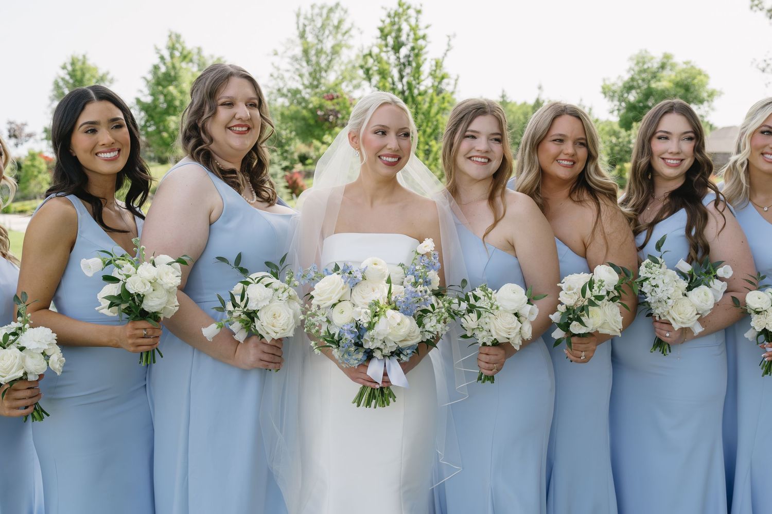 Bride in white gown with bridesmaids in light blue dresses holding white floral bouquets outdoors.