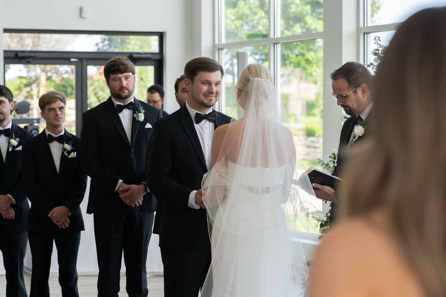 Groom smiling at bride during indoor wedding ceremony with groomsmen in black tuxedos standing nearby.