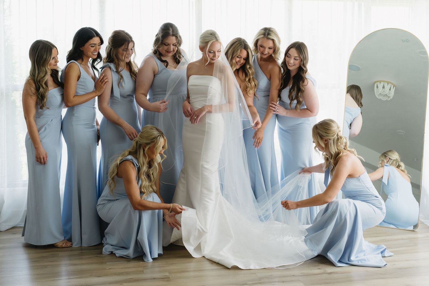 Bride in white gown surrounded by bridesmaids in blue dresses adjusting her veil and train indoors.