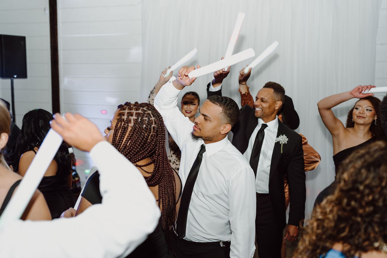 Party guests dance enthusiastically while holding glowing LED foam sticks at a wedding reception.