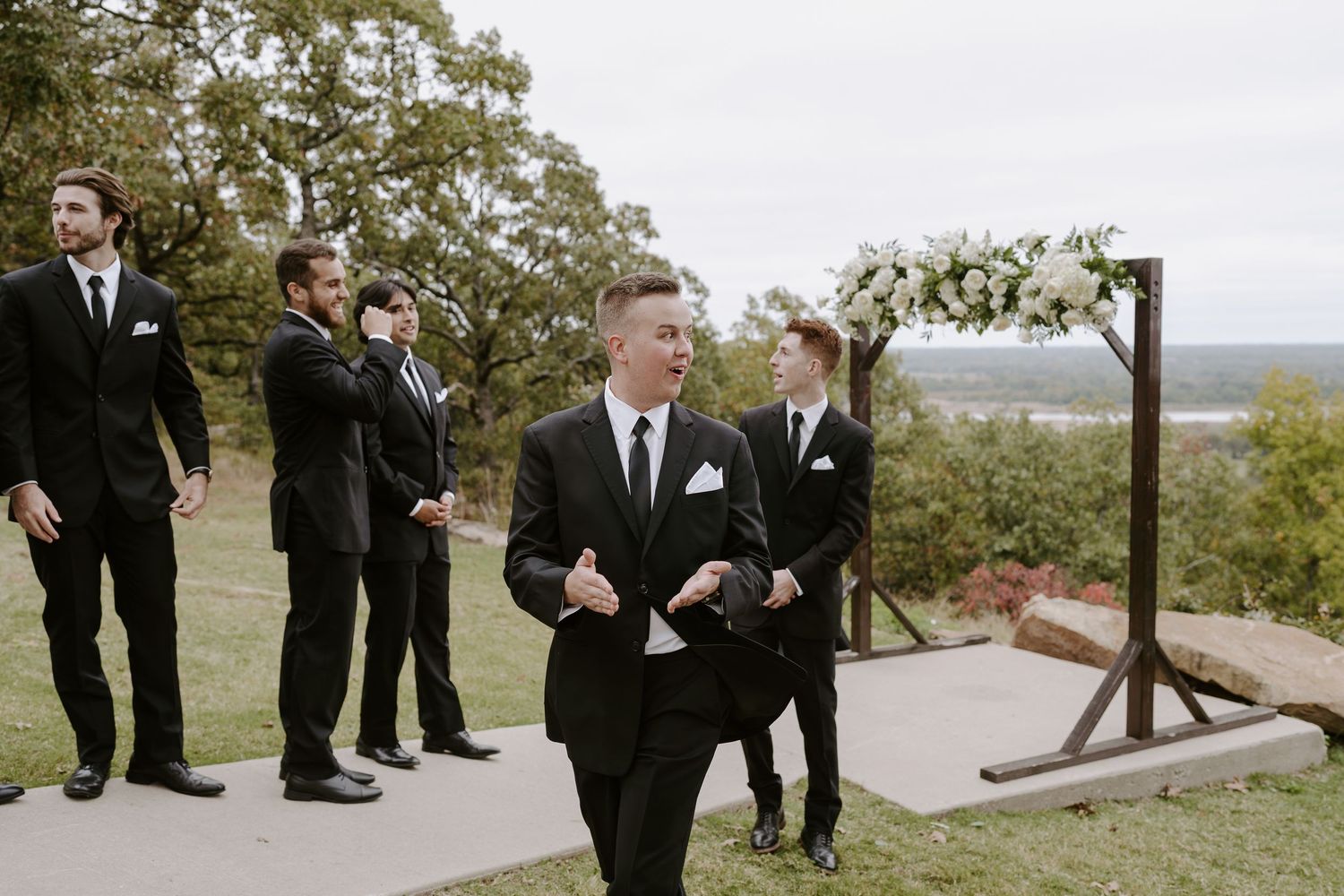 Wedding party members in black suits pose along wooden pathway outdoors.