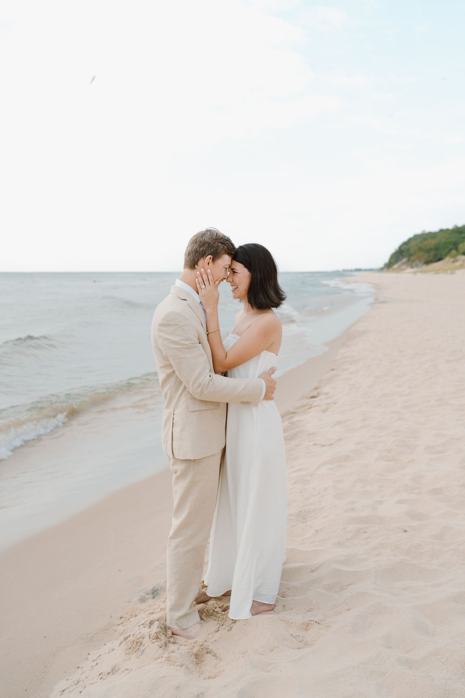 A couple shares a romantic embrace on a sandy beach with waves lapping at the shore.