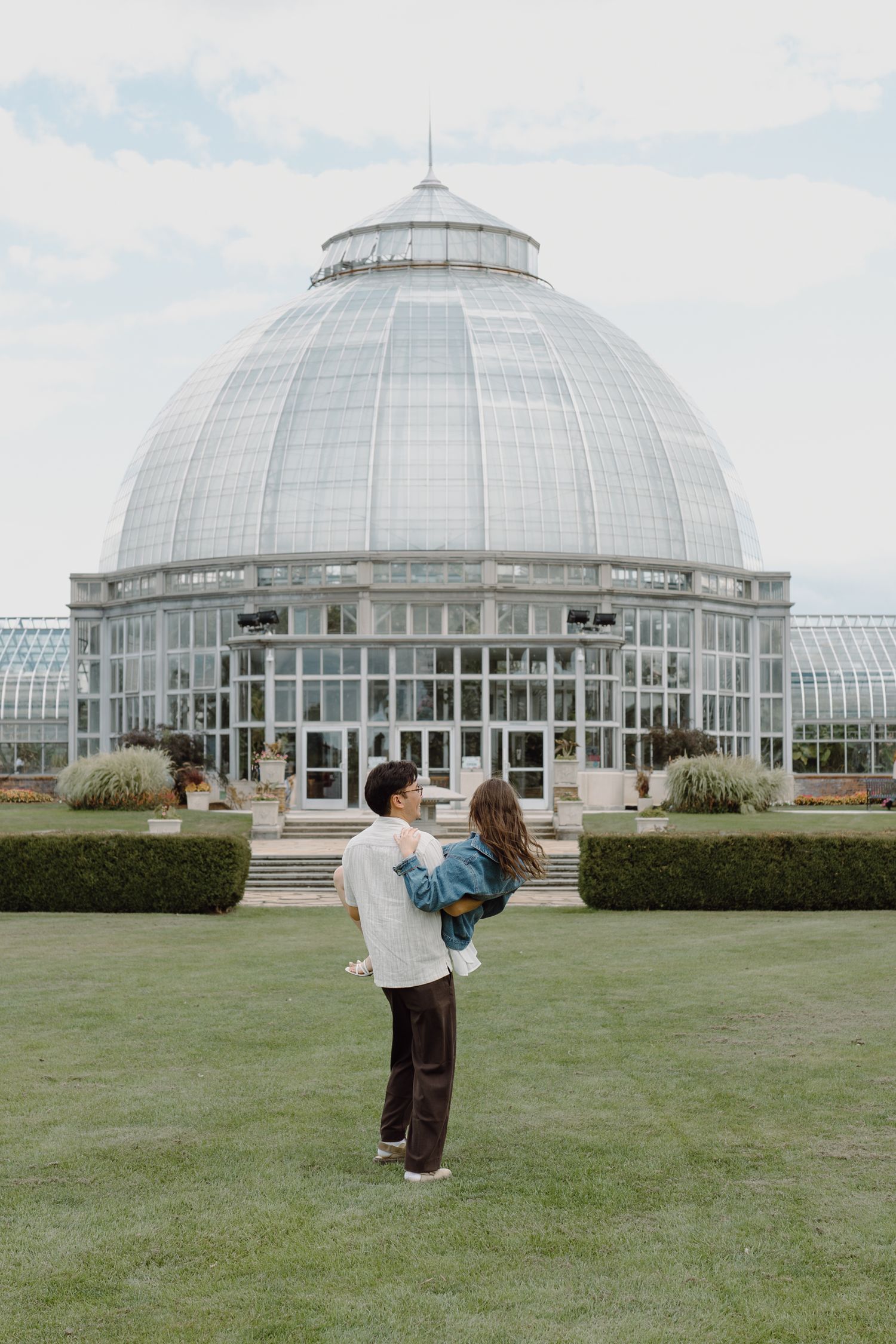 People dancing playfully on grass in front of an ornate glass conservatory building under blue skies.