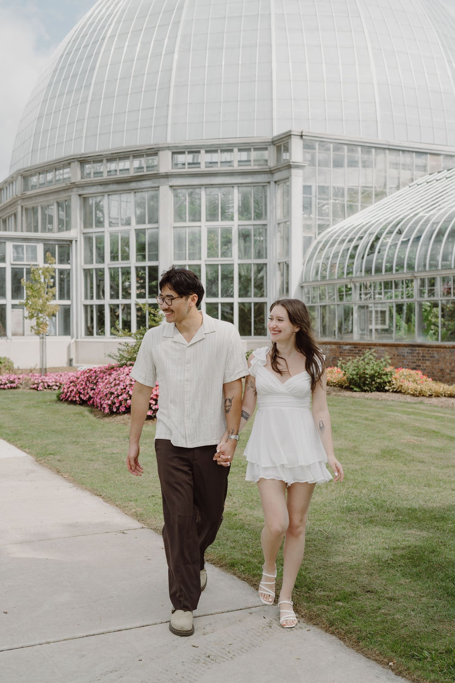 Two people stroll hand in hand along a path beside a large glass conservatory building.
