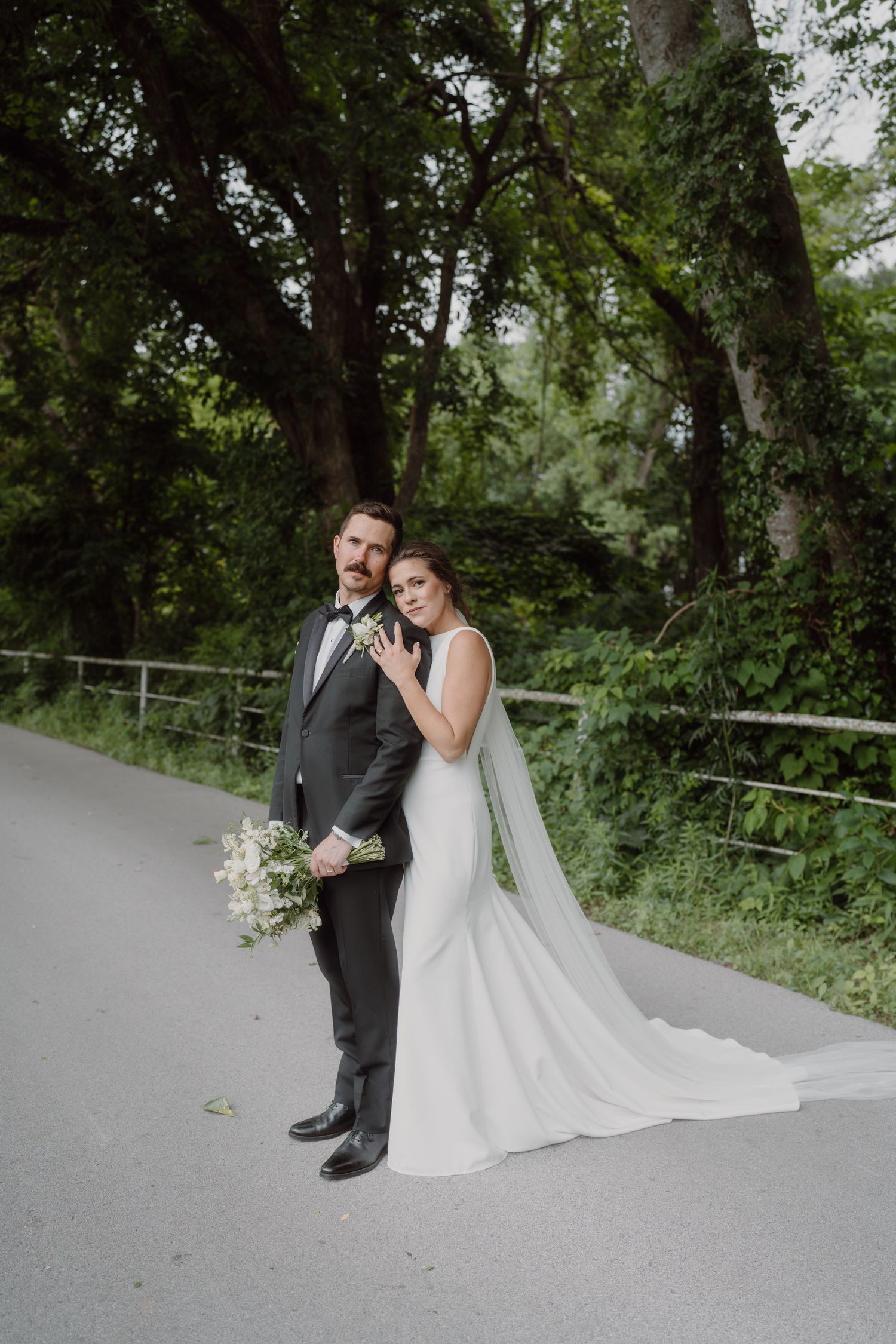 Newlyweds share an intimate moment on a paved forest path surrounded by lush greenery.