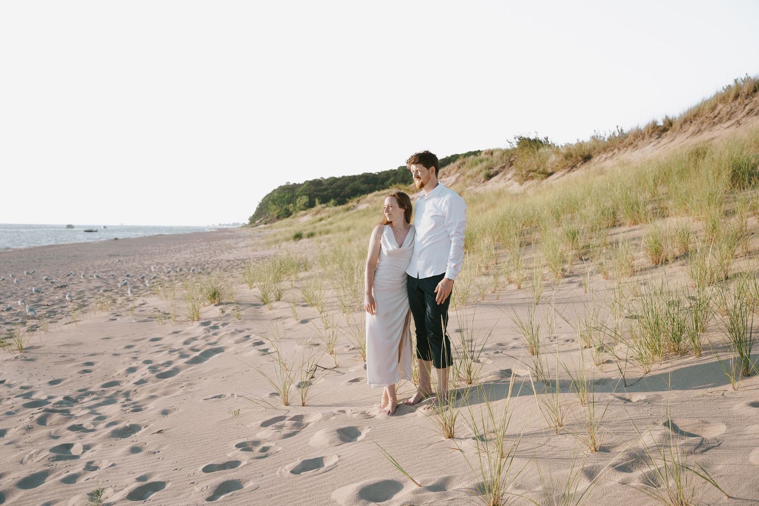 Two people pose together on sandy dunes overlooking a vast beach landscape.