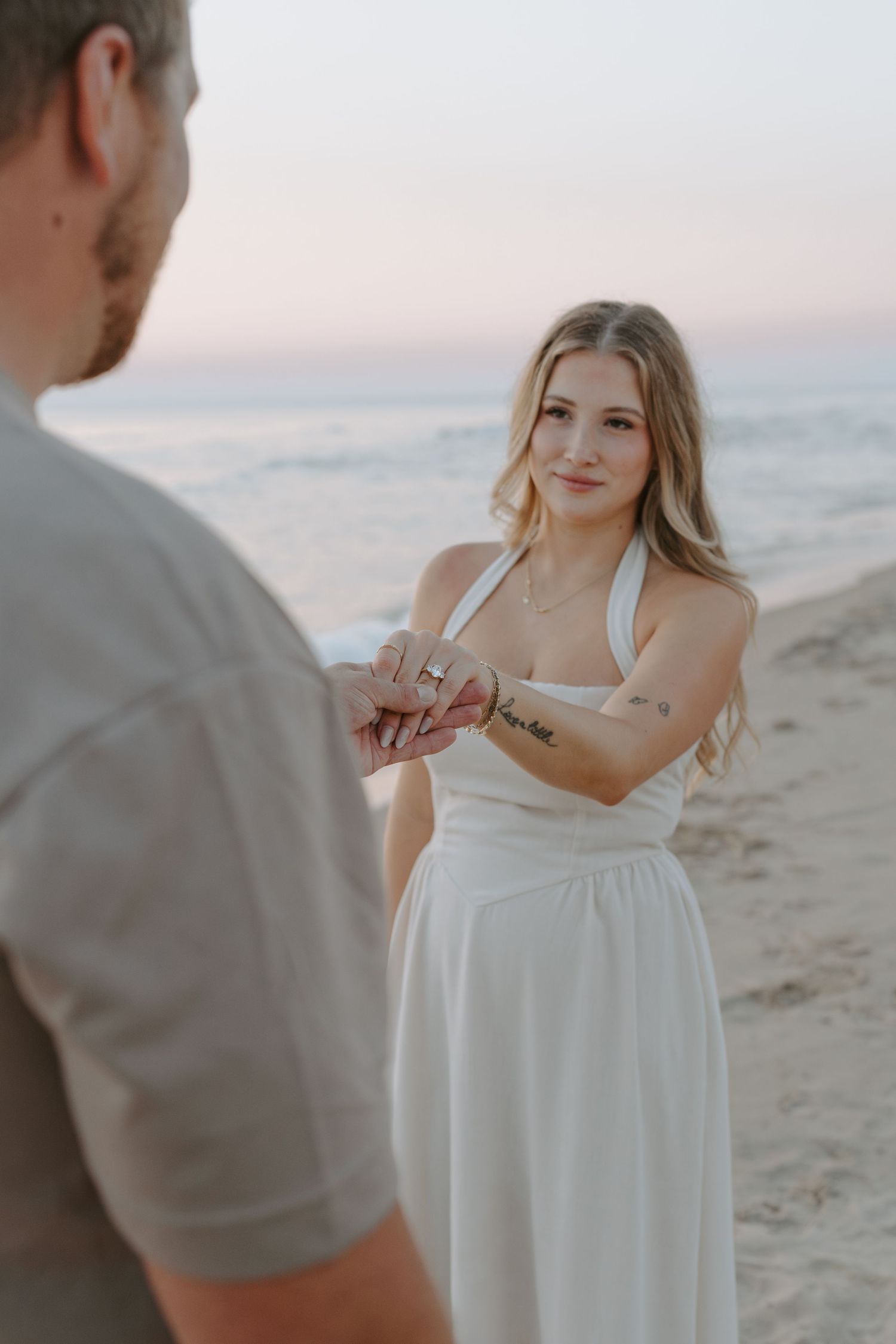Woman in white dress poses with engagement ring during beach proposal.
