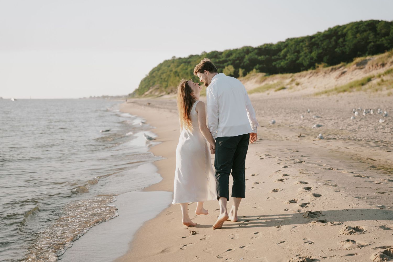 A couple strolls together on a sandy beach with waves and cliffs in the distance.