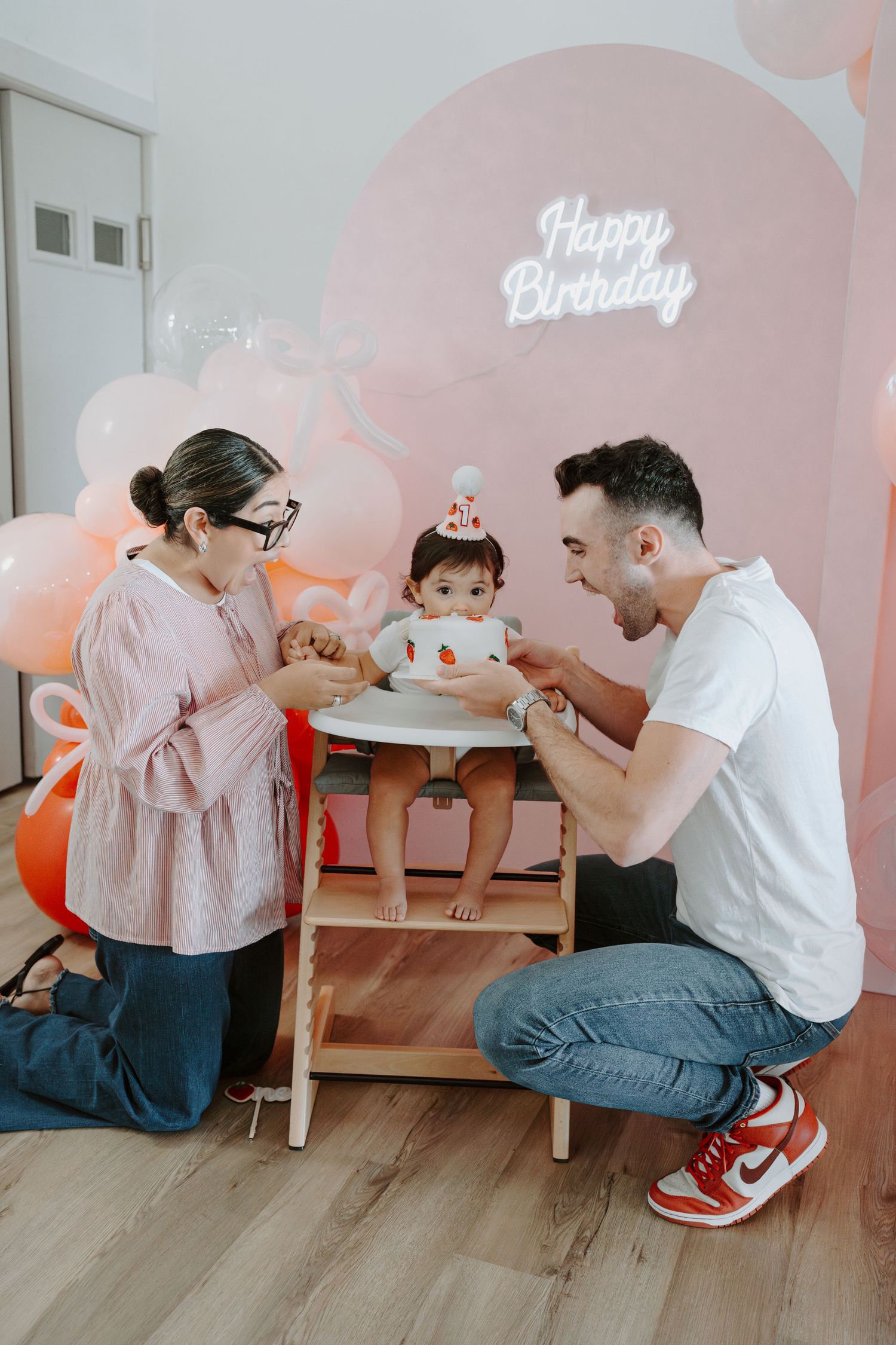 Family gathered around baby's high chair during birthday celebration with pink backdrop.