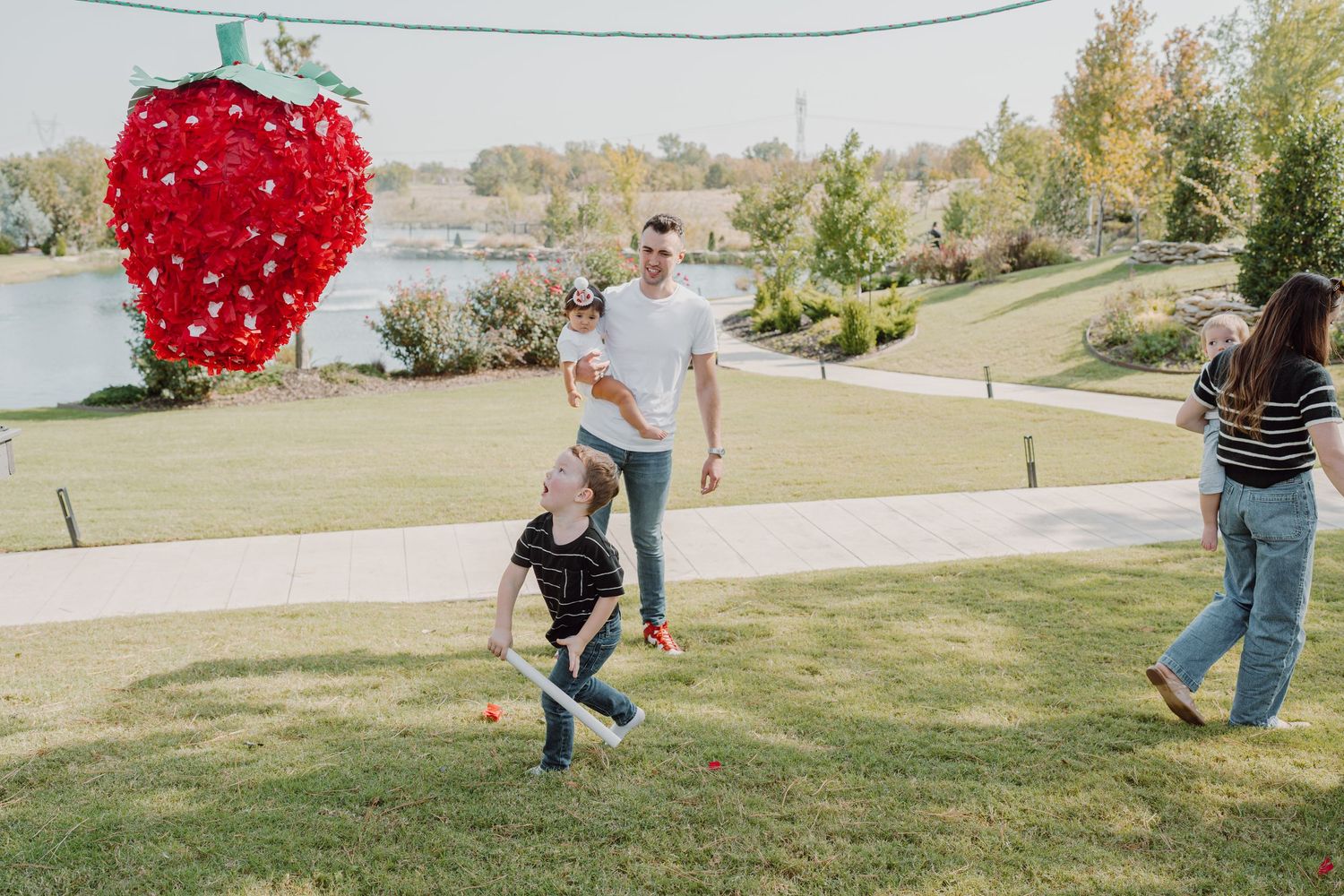 A large red heart-shaped piñata swings on a string against a grassy outdoor backdrop.