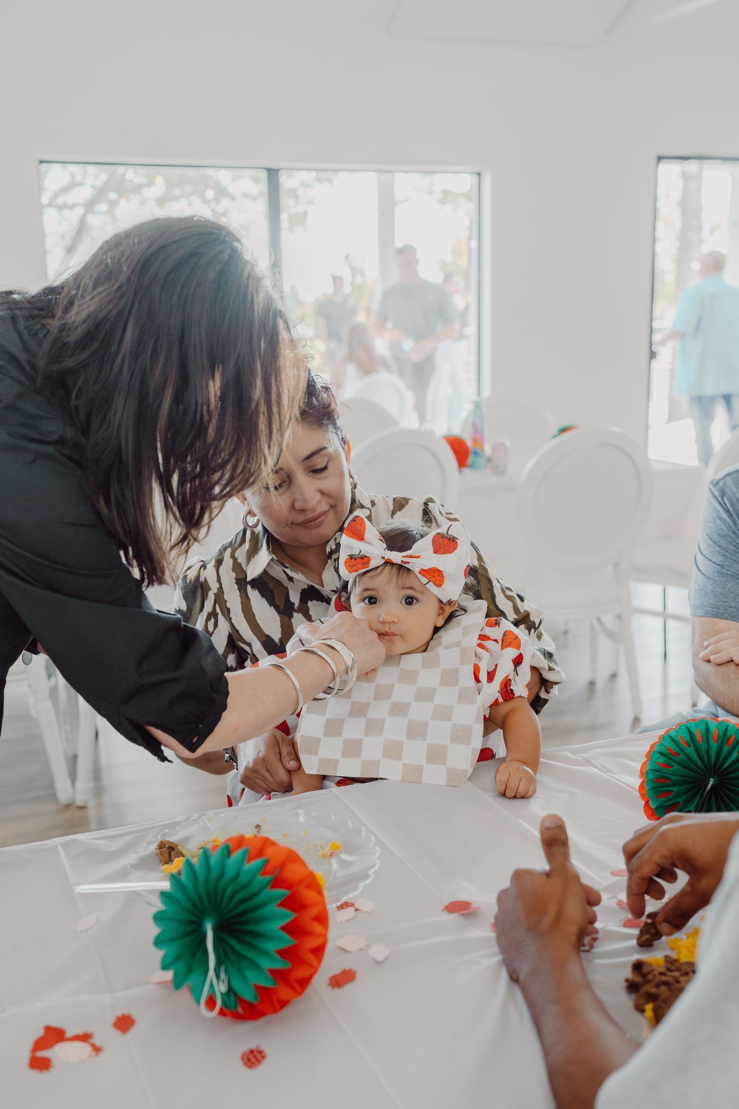 Baby reaching for items on white table during celebration with decorative green and orange paper fans.