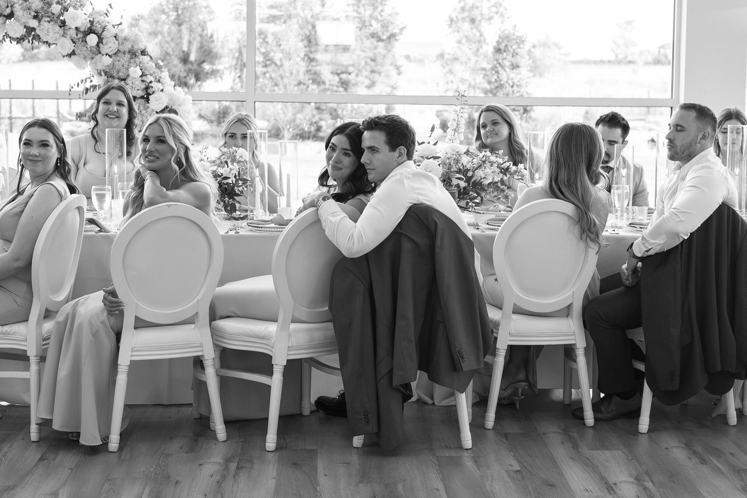 Black and white wedding reception photo of couple embracing at elegant dinner table surrounded by guests.