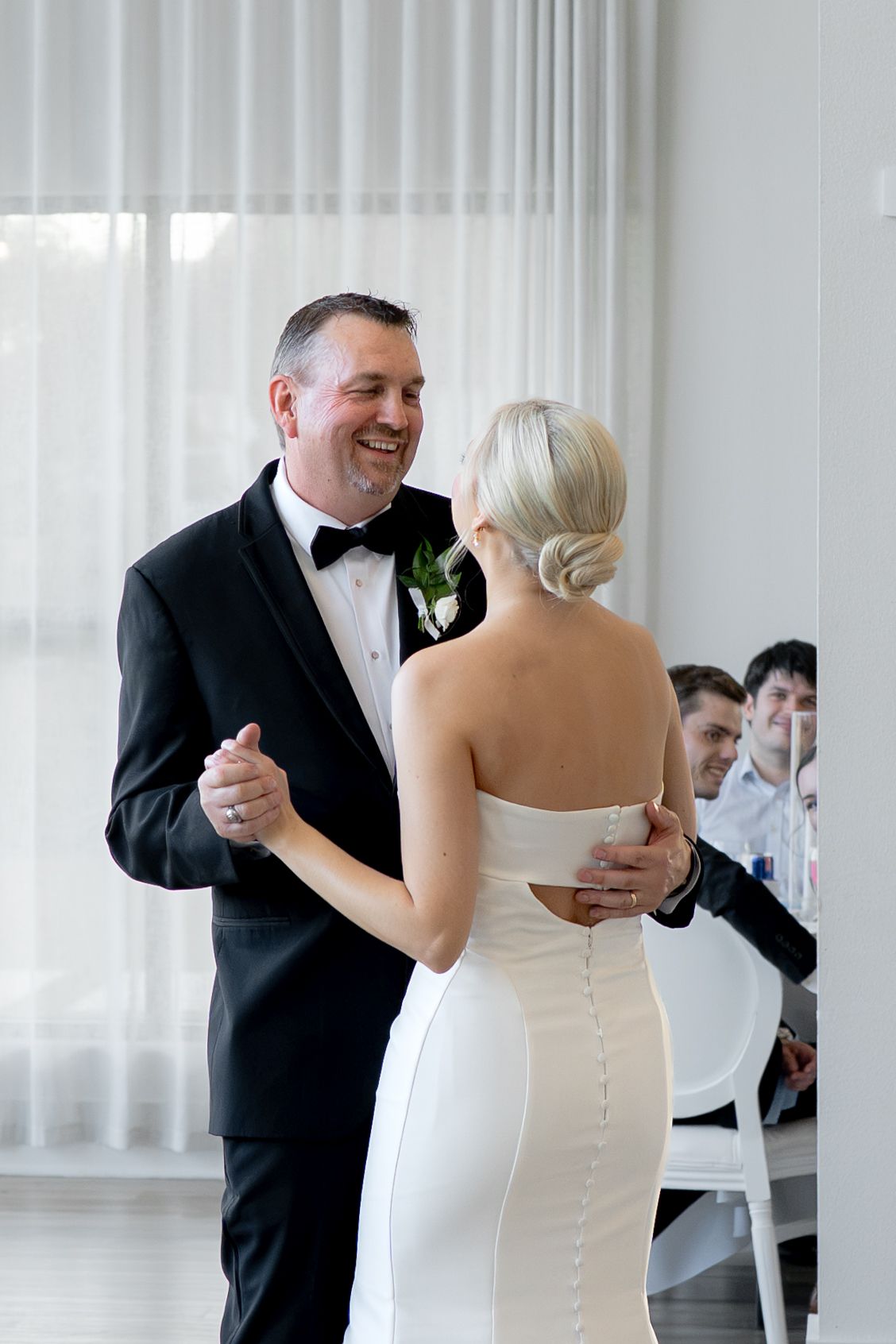 A bride and groom share their first dance, smiling joyfully, with guests watching in the background.