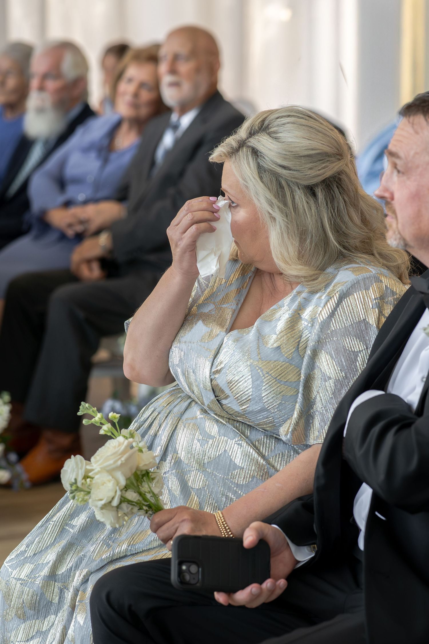 Emotional pregnant woman in silver dress dabbing tears at wedding ceremony while holding white flowers.